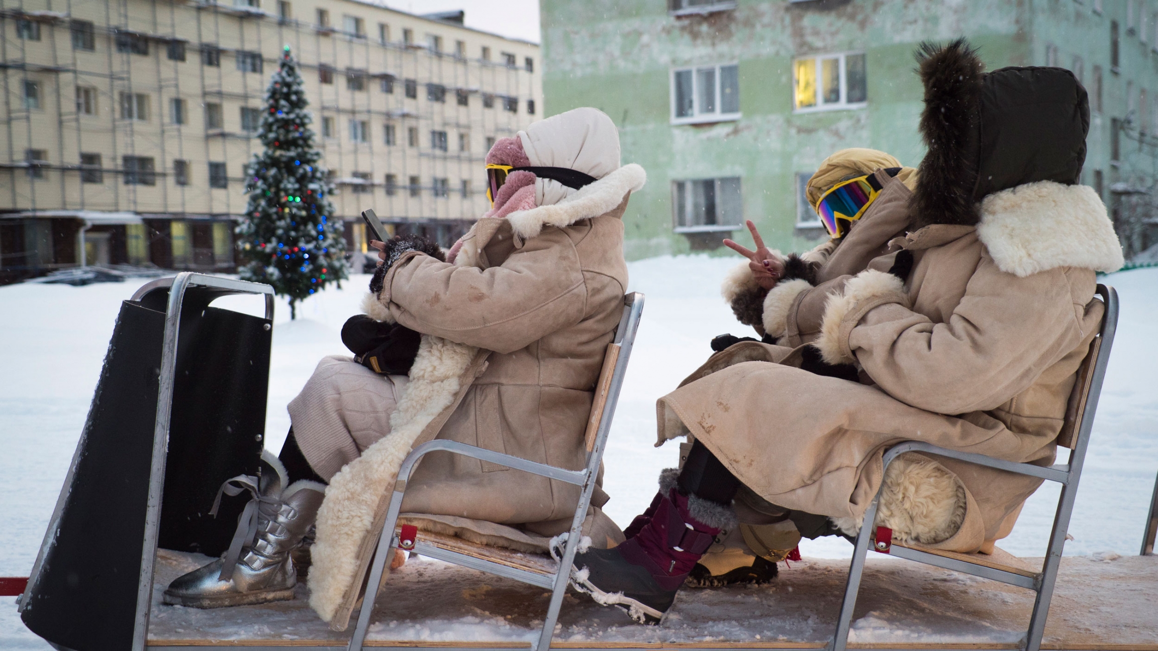 Tourists from China sit in a snowmobile trailer in Teriberka, Russia. A group of people are shown wearing large jackets and sitting in two rows of seats in a trailer.