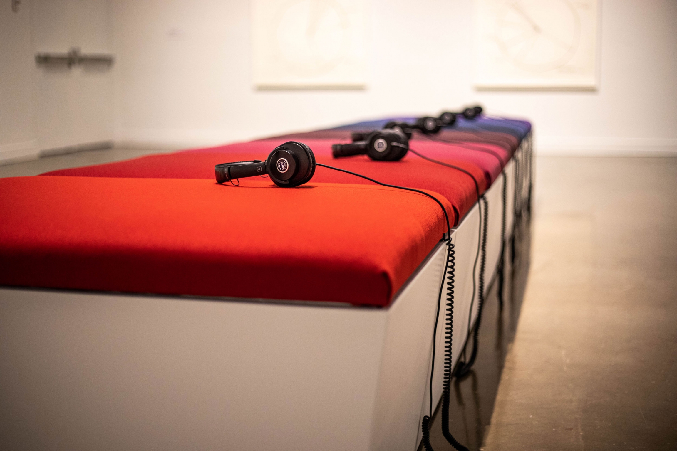 Artist Christine Sun Kim's audio installation entitled, “lullabies for roux (2018)." A long bench is shown with brightly colored cushions and headphones on each cushion.