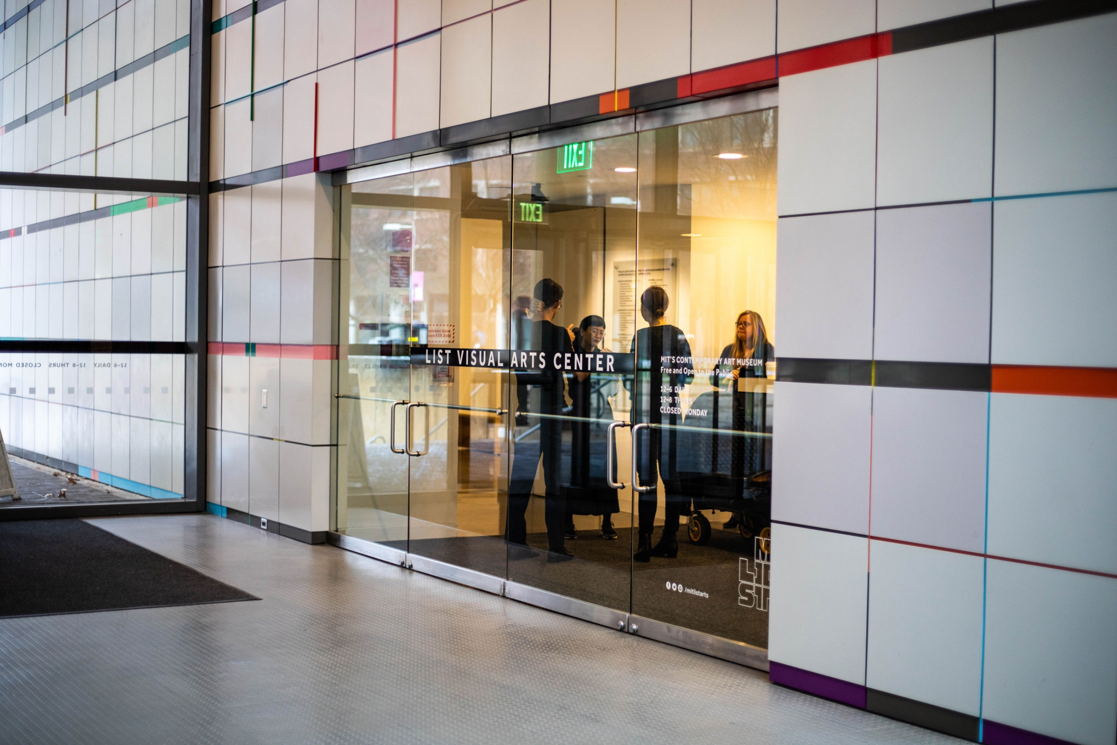 Christine Sun Kim (C) at the MIT List Visual Arts Center where her current artwork, "Off the Charts," is being exhibited. Several people are shown through glass doors with the words, 'List Visual Arts Center,' printed on them.