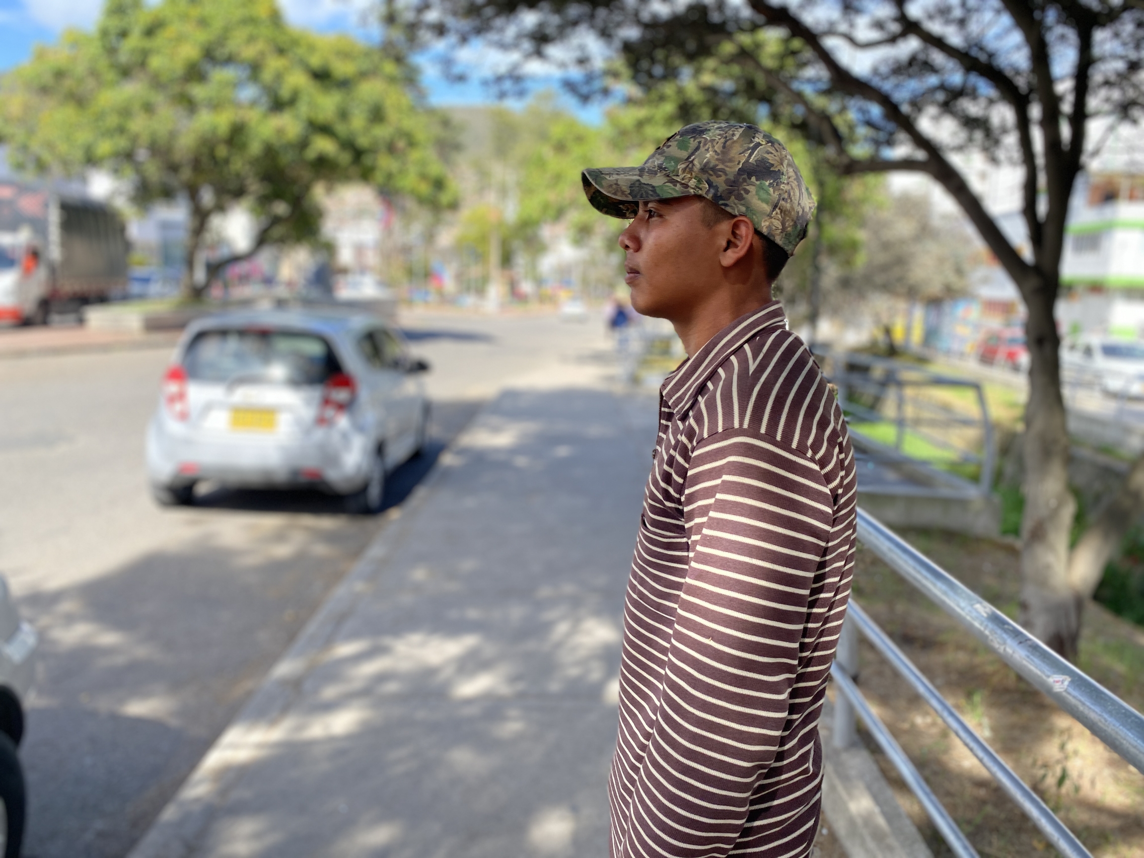 Alexander Tiapa Maita, a former Venezuelan soldier who defected in April last year, takes a break in Pamplona, Colombia where he works as a gardener. A young man wears a camouflage hat with brown and cream striped shirt and stands outside near a parked car.