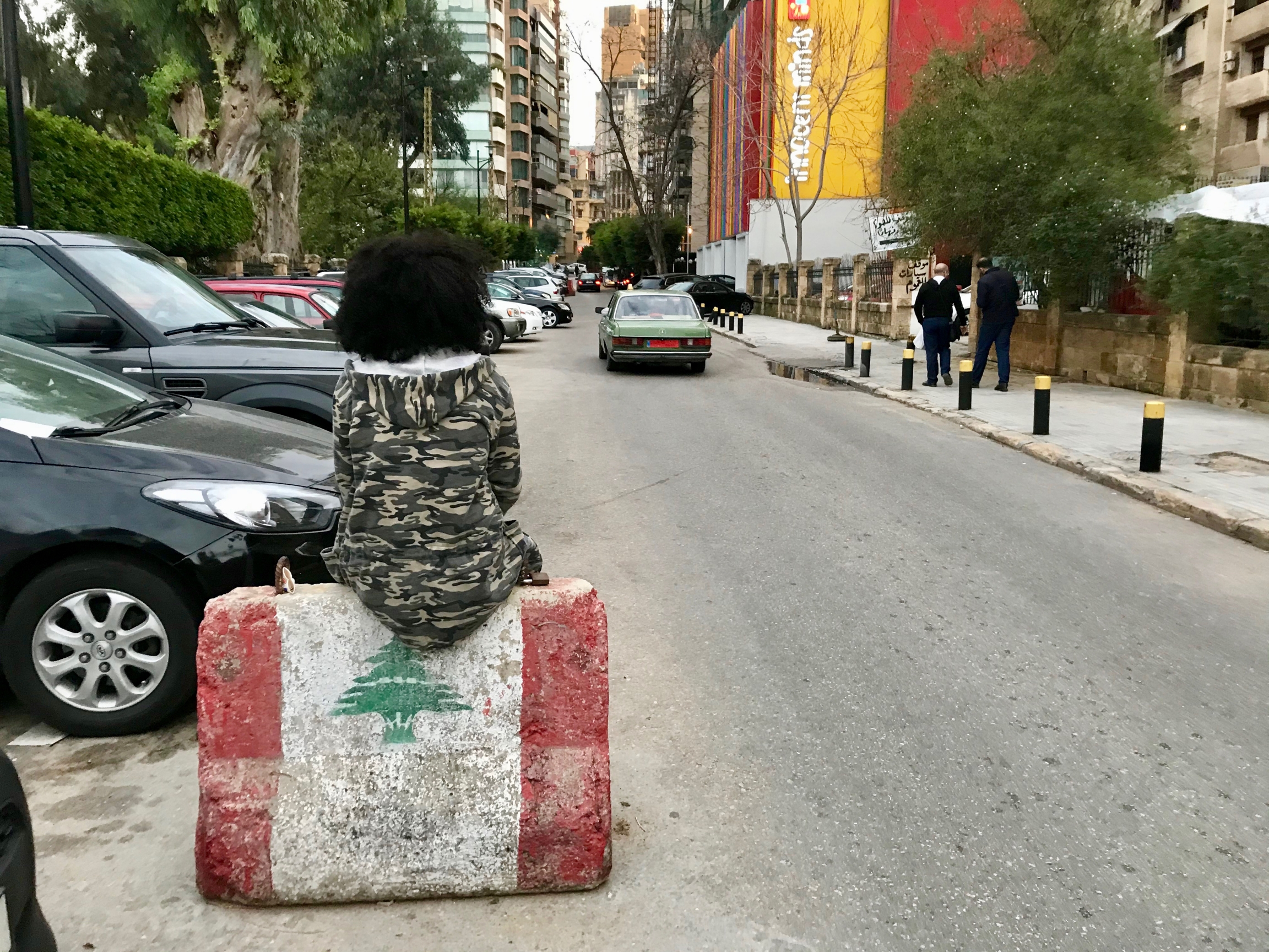 Tsigereda perches on a barricade painted with the Lebanese flag in the Hamra neighborhood of Beirut, Lebanon. A young woman wearing a camouflage jacket sits on a barricade in middle of street.