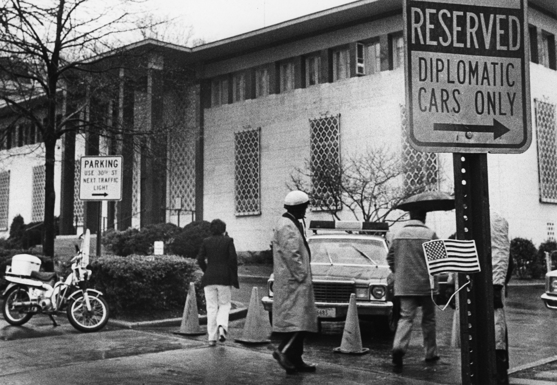 The exterior of the Iranian Embassy as workers got the word to pack their bags and go home, in Washington, DC, Dec. 13, 1979. Several people and a police car are shown in this black and white photograph with the former Embassy of Iran in the background.