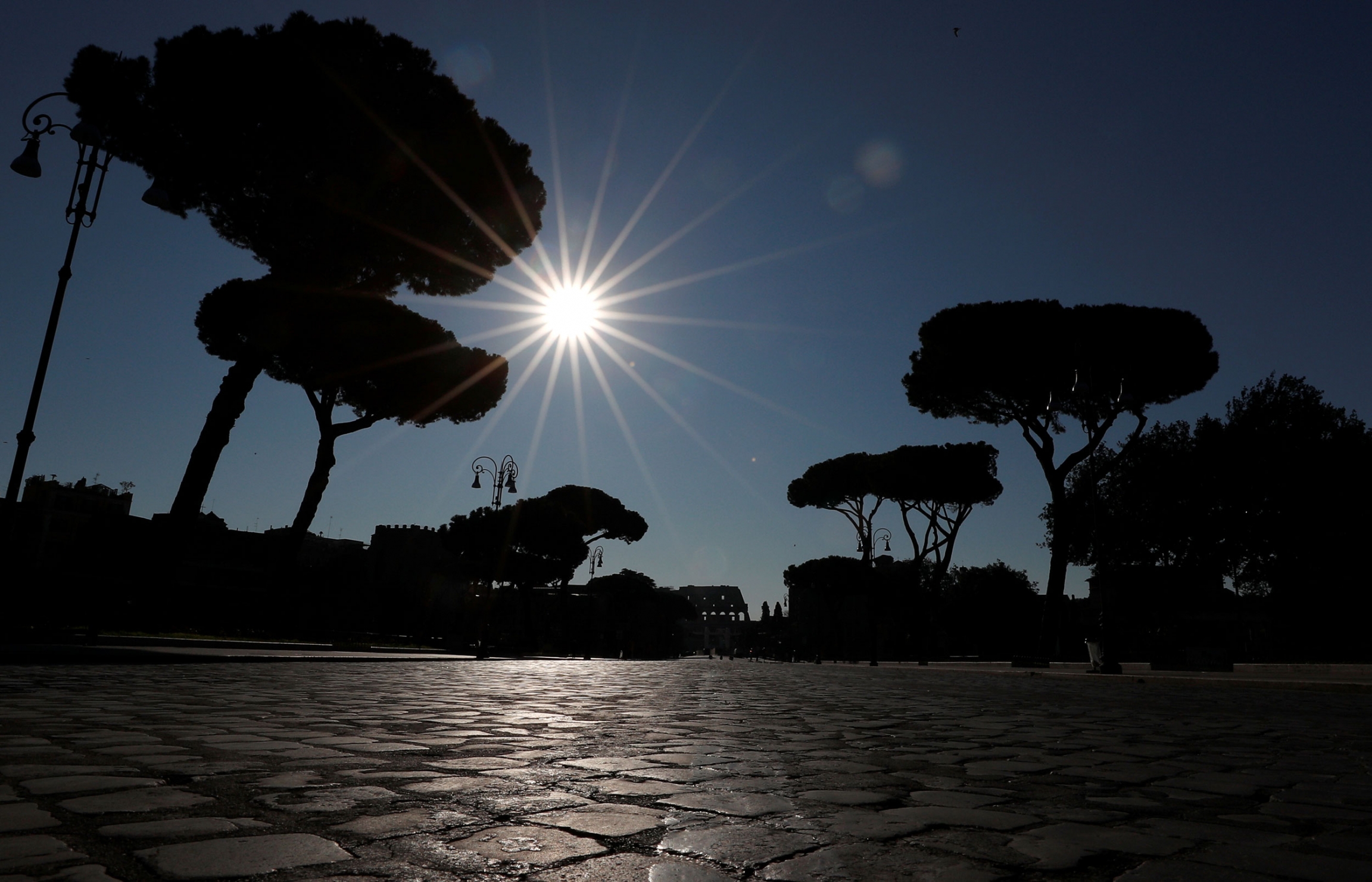 A virtually deserted street near the Colosseum is pictured after a decree orders for the whole of Italy to be on lockdown in move to contain the coronavirus, in Rome, Italy, March 10, 2020. A bright sun shines in a blue sky with several trees shown in shadow.