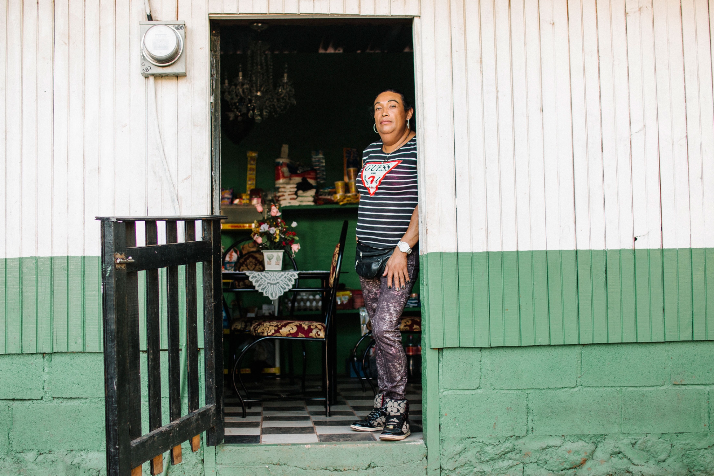 Talia, a former Cattrachas activist, say she was brutalized by the police and military as a trans sex worker. These days is focused on staying safe and running her business: a small convenience store she operates out of her home. Talia is shown standing in the entryway of her convenience store that has a dark colored, slated wooden gate.