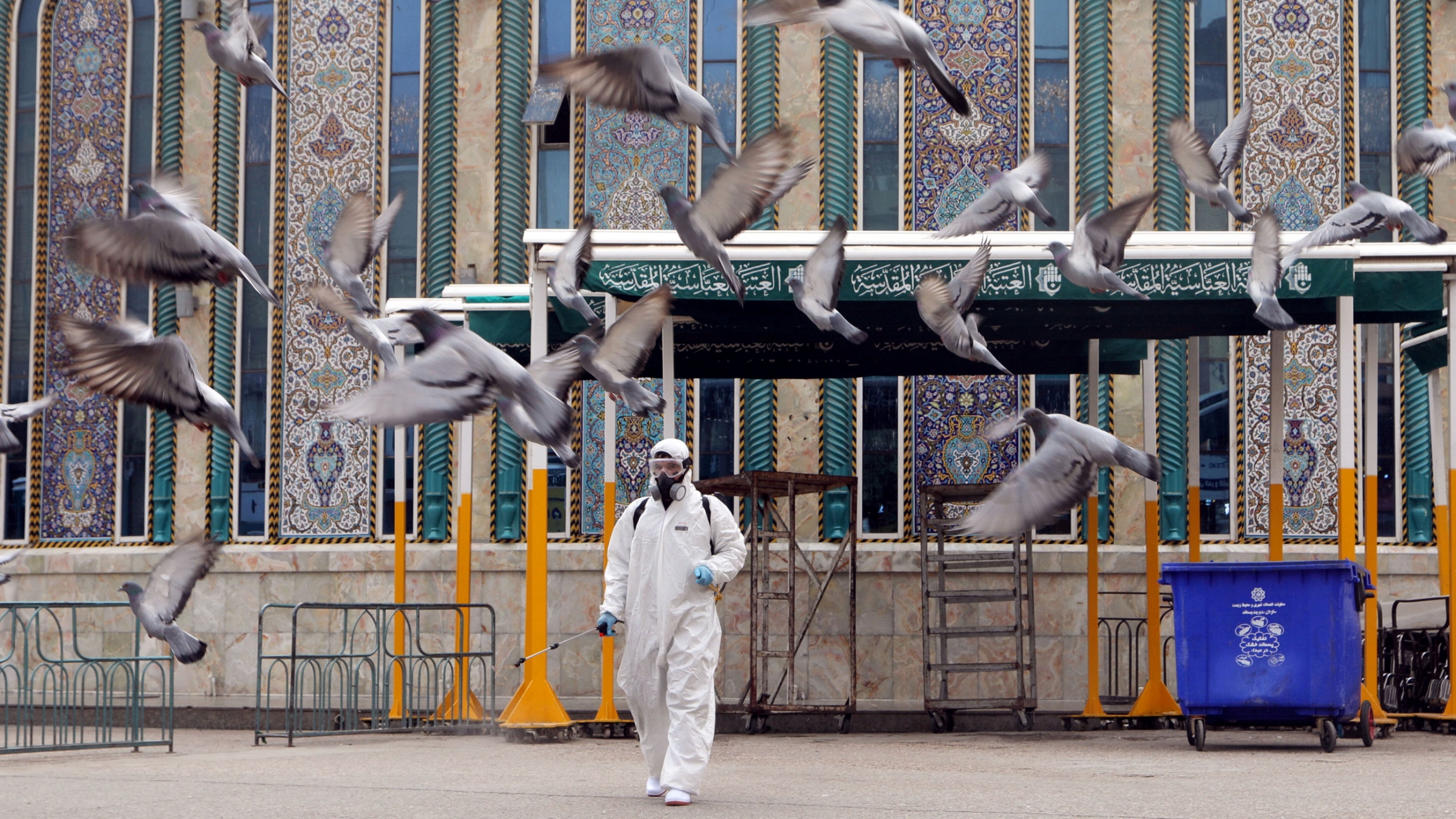 A worker in a protective suit sprays disinfectants near Imam Abbas shrine as a precaution against the coronavirus, following the outbreak, in the holy city of Kerbala, Iraq, March 15, 2020. A man is shown wearing a full body protective suit, spraying disinfectant as pigeons fly away.