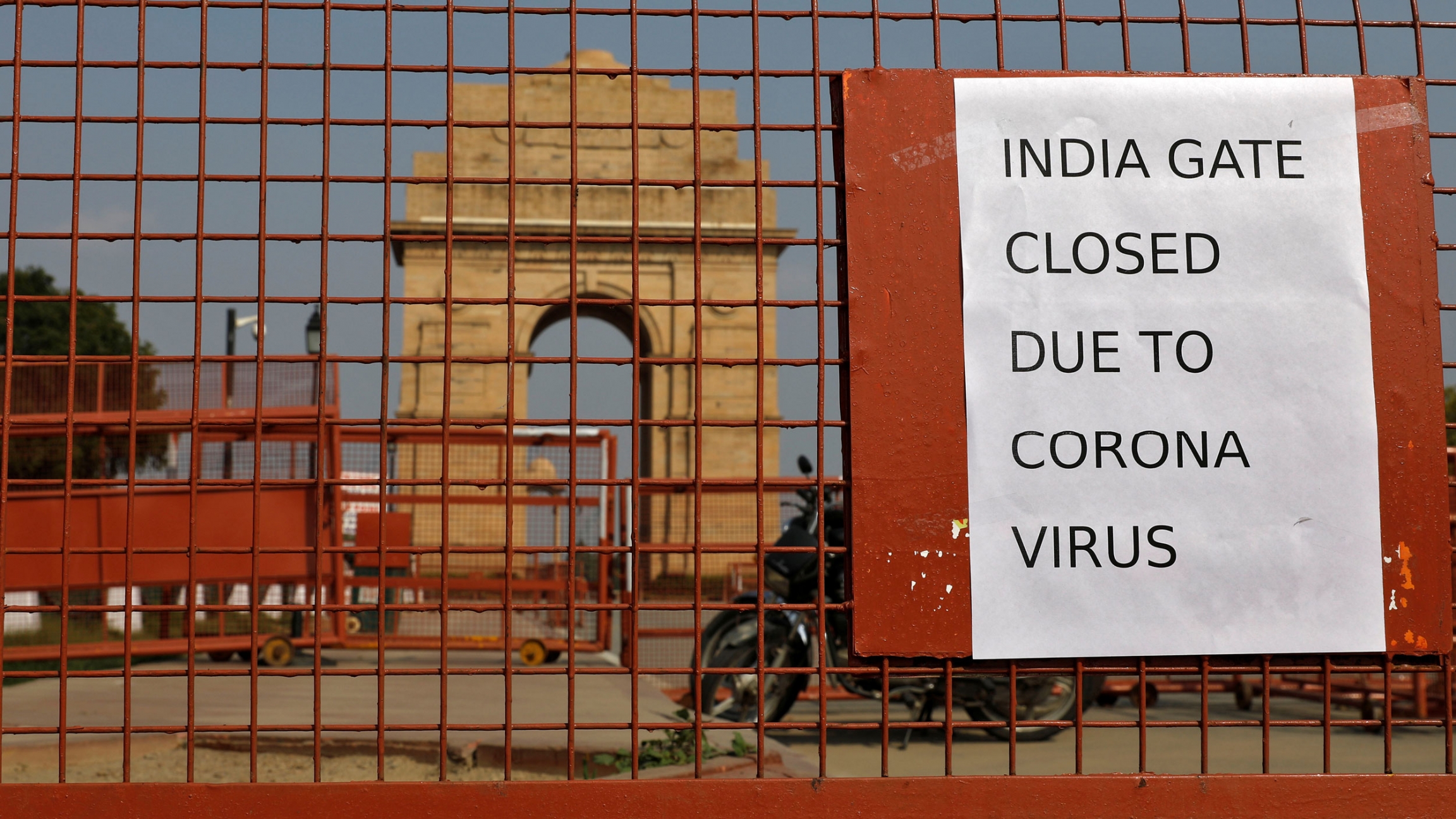 A sign pasted on a security barricade is seen after the India Gate war memorial was closed for visitors amid measures for coronavirus prevention in New Delhi, India, March 19, 2020. A close-up photograph of a reddish metal gate with a motorcycle parked behind it and the India Gate war memorial is shown off in the distance.