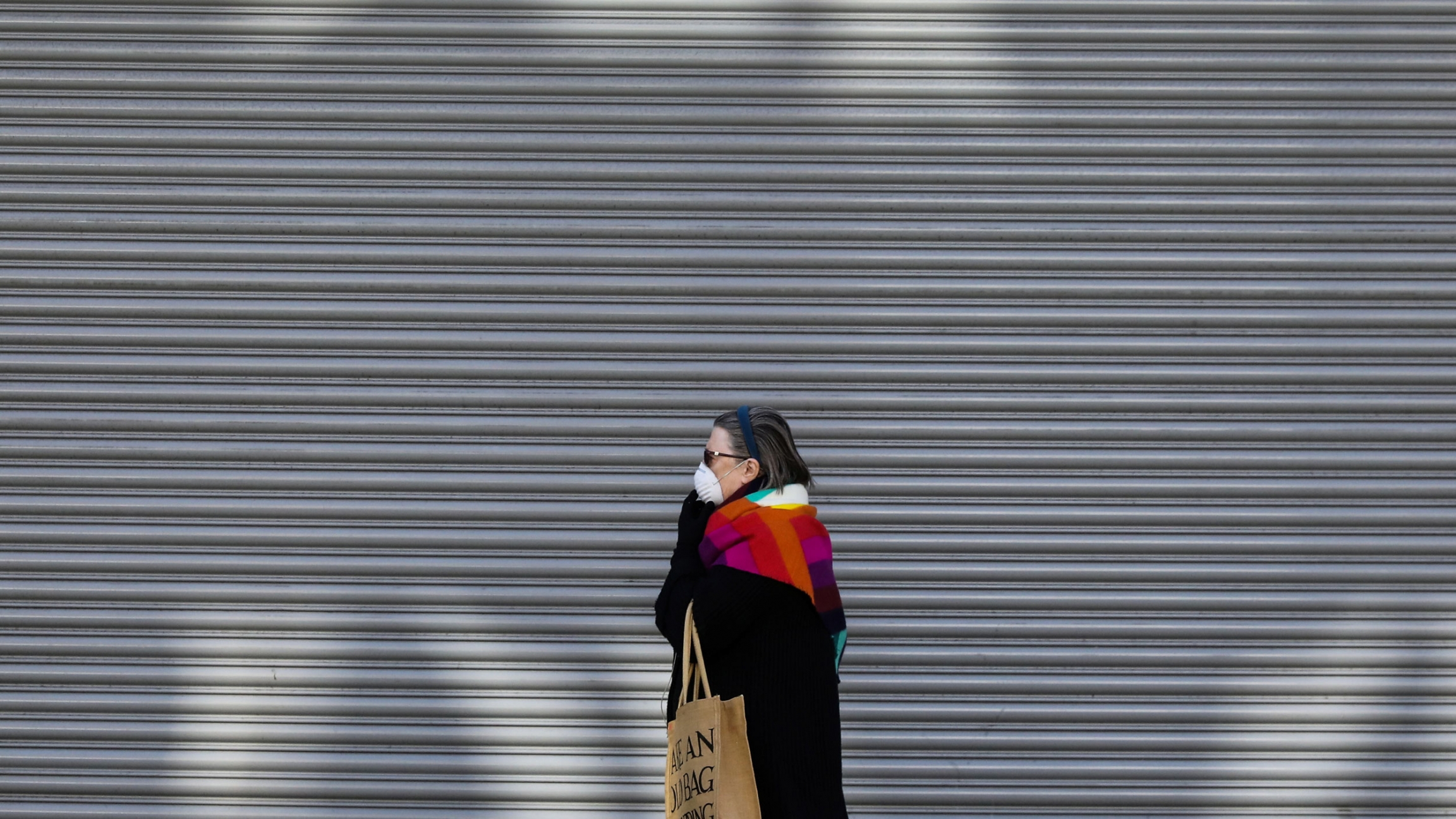 A woman walks past a shuttered shop as the spread of the coronavirus disease continues in London, Britain, March 24, 2020. A older woman is shown holding a mask to her face as she walks past a shop with a metal gate pulled down.