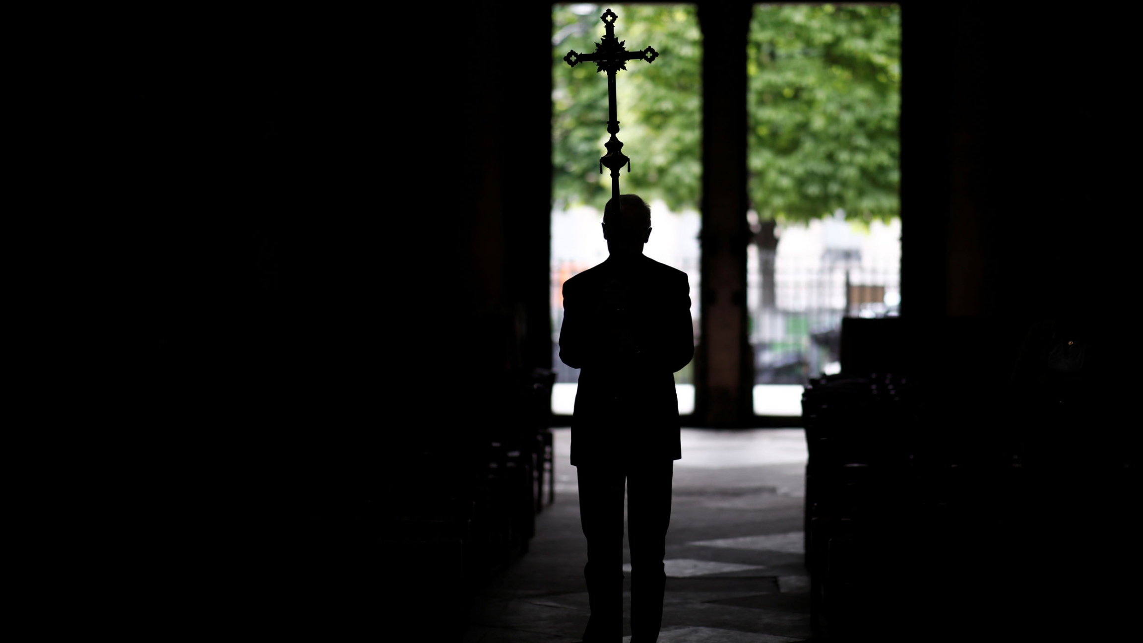 A man is shown in shadow standing in an open area and holding a cross above his heads.