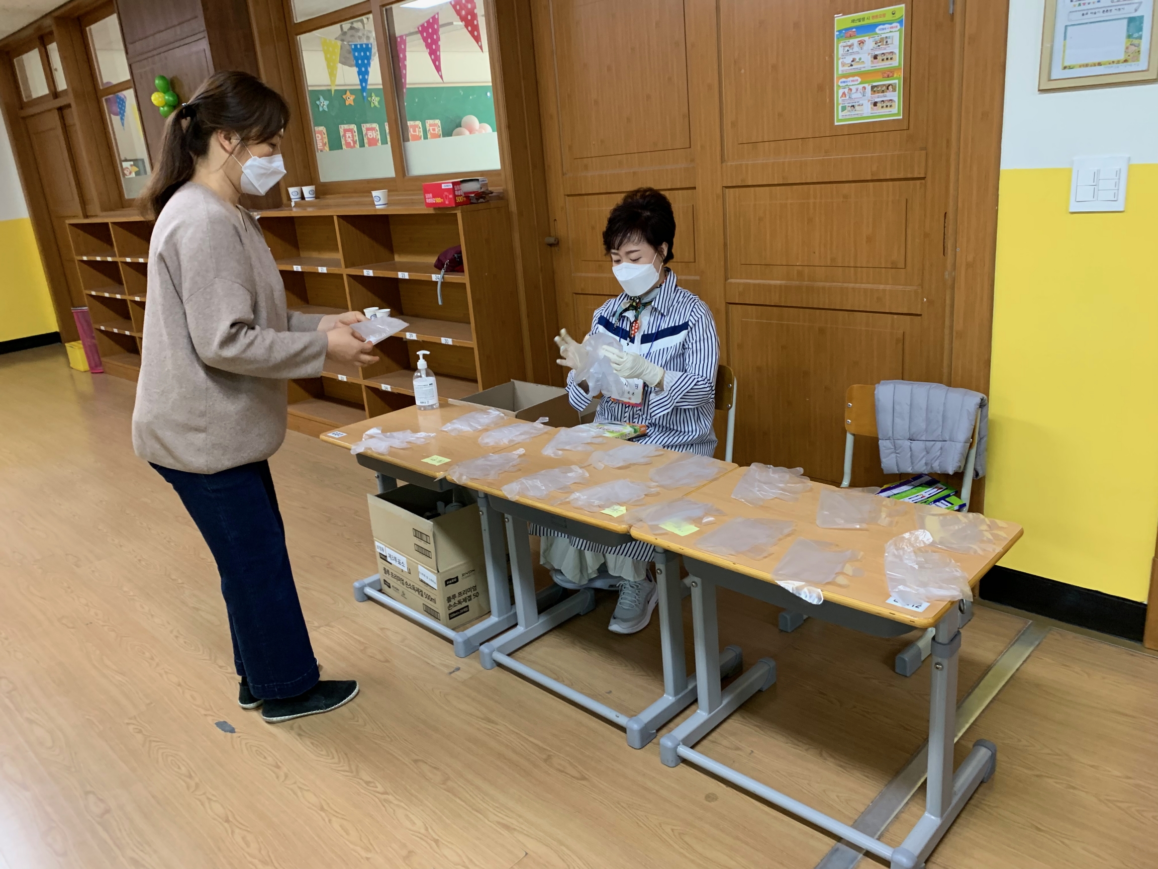 At a polling station, two people prepare gloves for voters casting ballots amid the coronavirus outbreak in South Korea. One person wears a mask and sits at a table facing another person wearing a mask and gloves.