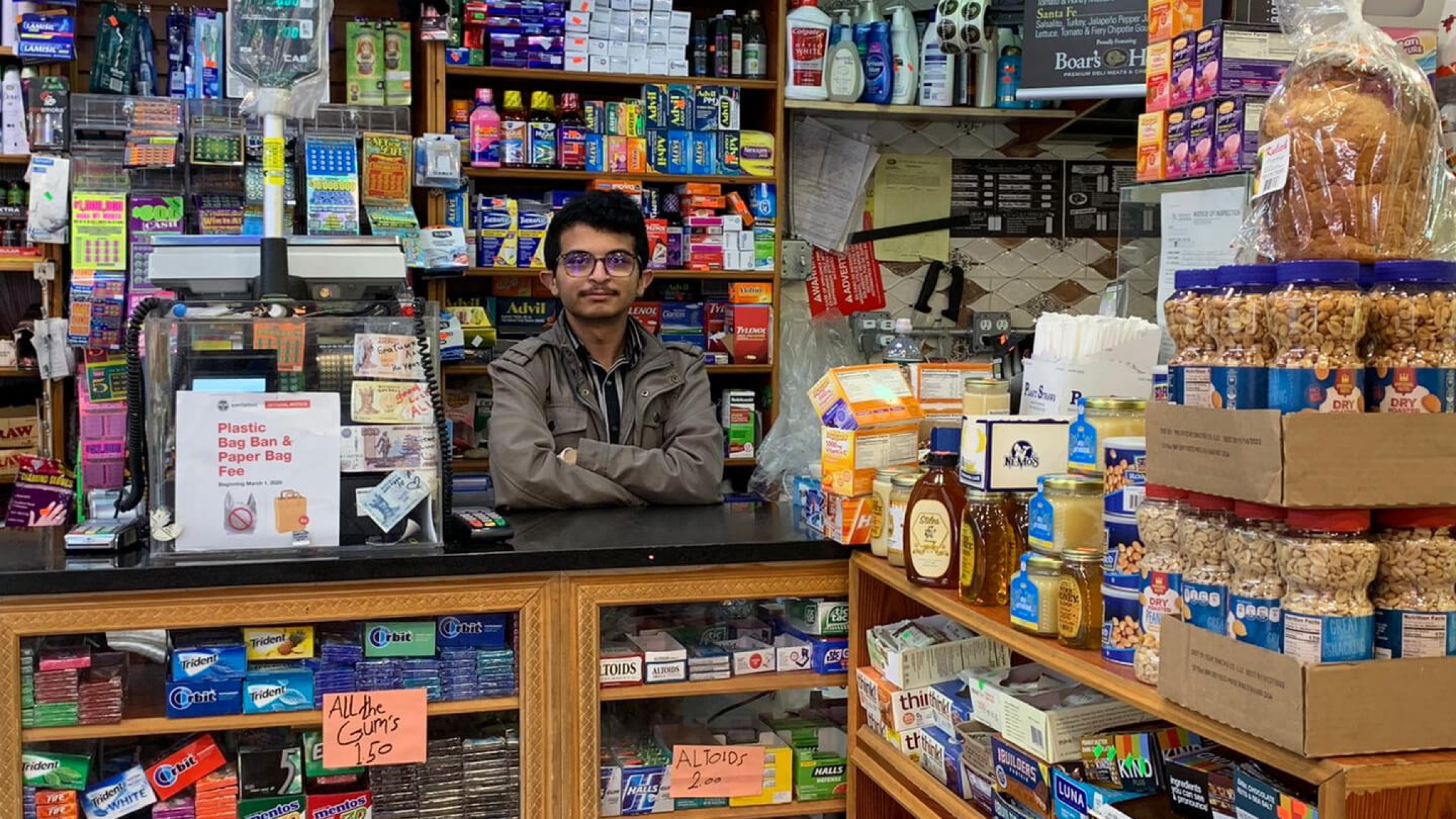 A young man wearing glasses stands inside his bodega filled with products in Brooklyn, New York.ย Nasim Almuntaser stands inside his packed bodega filled with products.