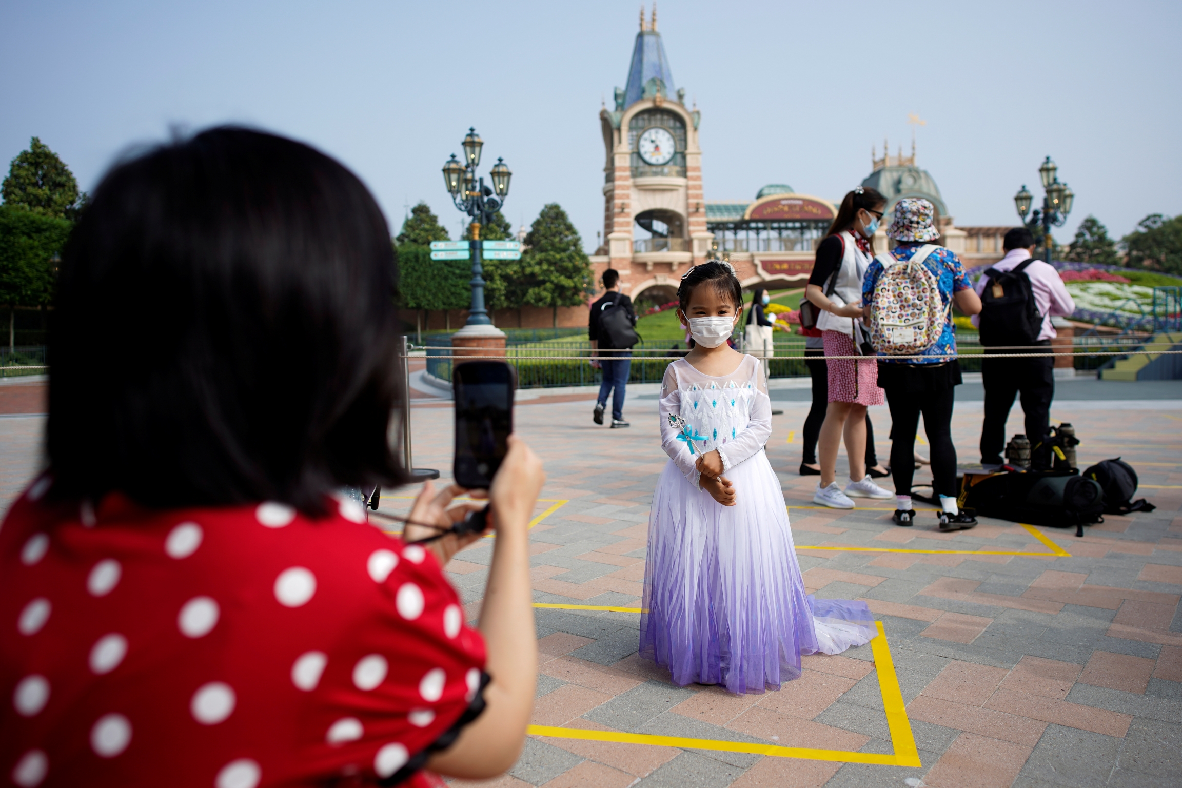 A girl wearing face mask poses for a picture at Shanghai Disney Resort as the Shanghai Disneyland theme park reopens following a shutdown due to the coronavirus disease (COVID-19) outbreak, in Shanghai, China, May 11, 2020. A mother photographs her child wearing a face mask and a princess gown at Disneyland in Shanghai.