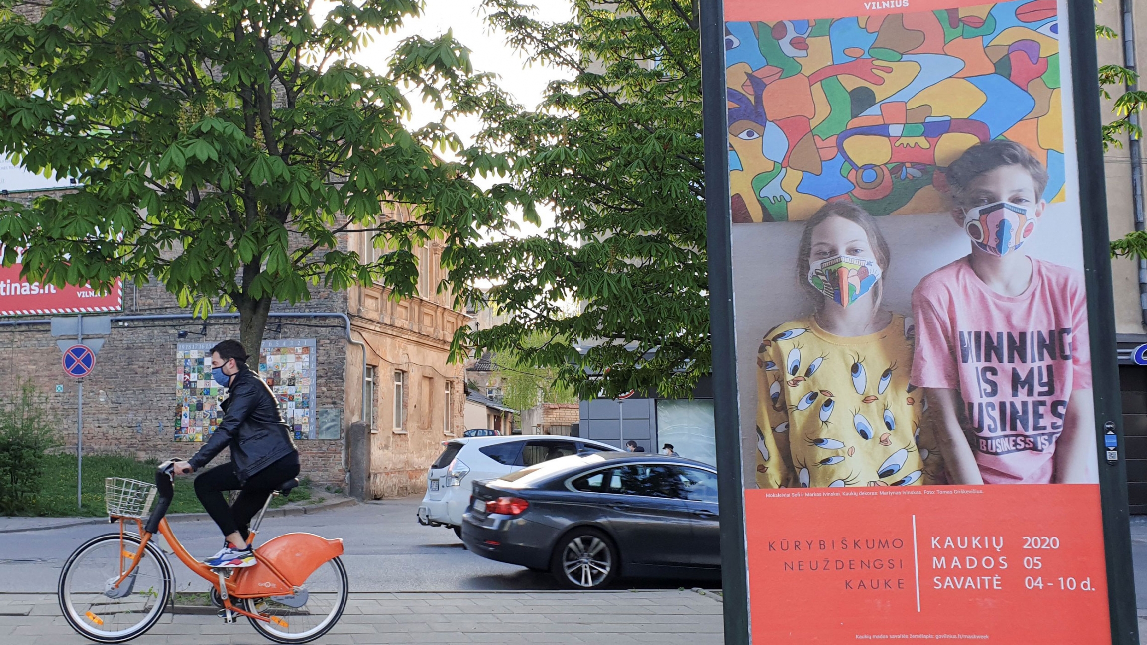 A bicyclist rides next to a billboard, a part of a "Mask Fashion Week" during the coronavirus disease (COVID-19) outbreak in Vilnius, Lithuania, May 5, 2020. A person on an orange bicycle rides past a billboard of people with masks