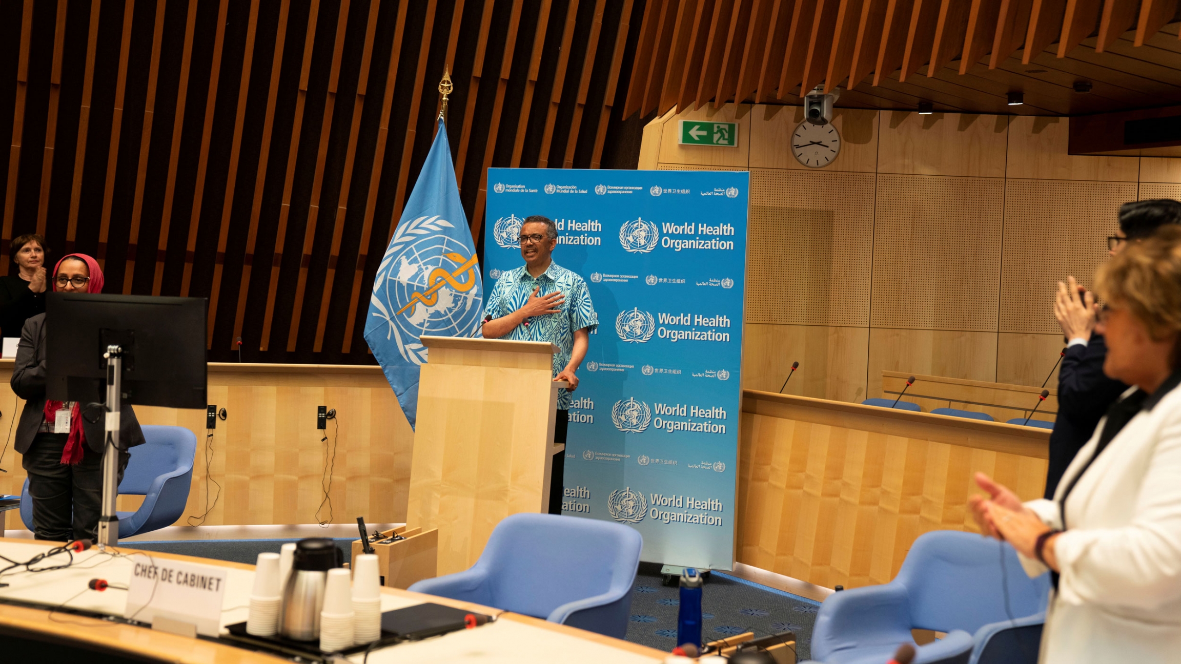 Tedros Adhanom Ghebreyesus, director-general of the World Health Organization attends the virtual 73rd World Health Assembly in Geneva, Switzerland, May 19, 2020. Tedros Adhanom Ghebreyesus is shown standing at a wooden podium with the blue WHO flag next to him and wearing a blue-print button down short-sleeved shirt.