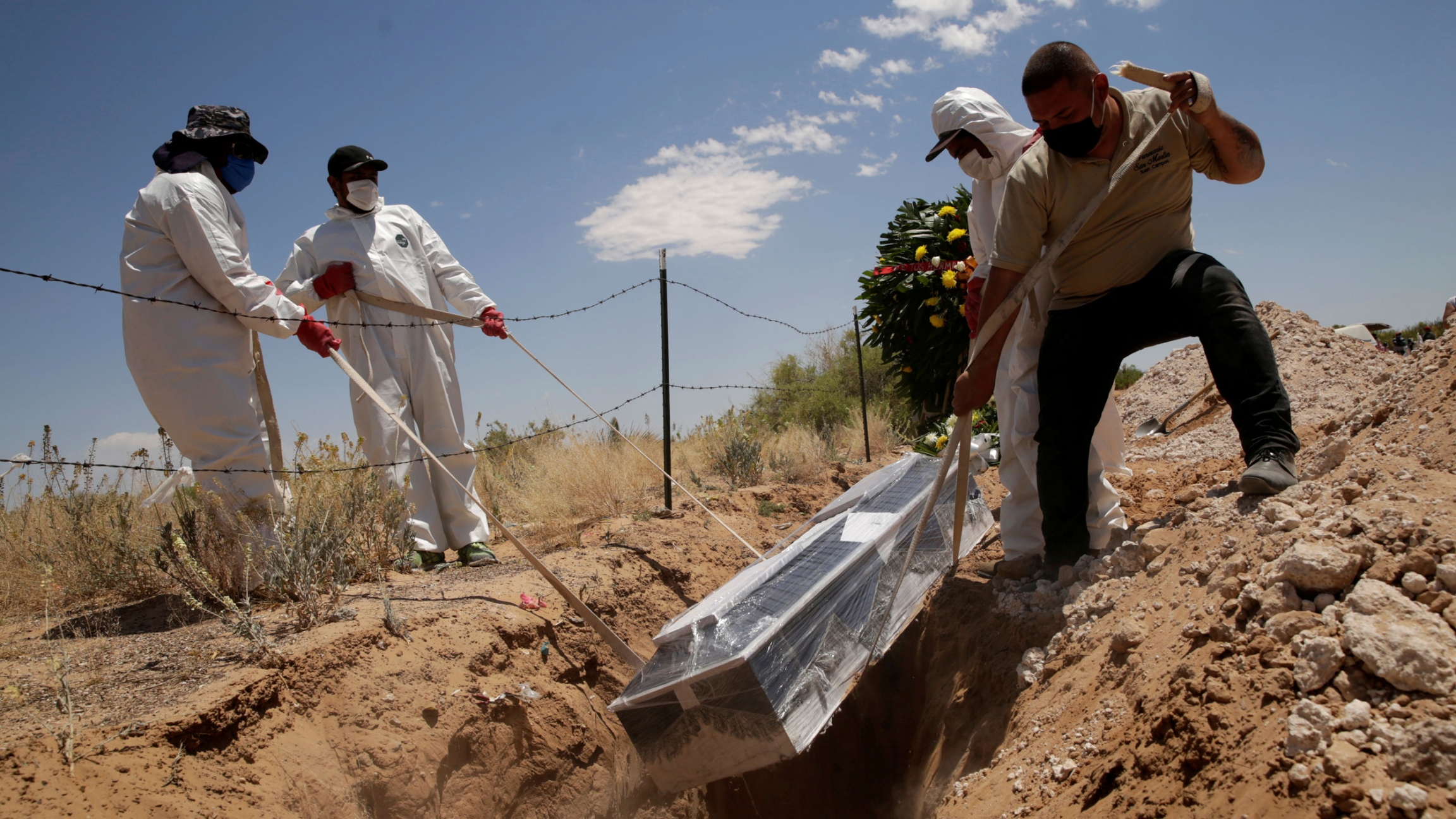 Cemetery workers place a coffin, containing the body of a 52-year old woman who died of the coronavirus disease, into a grave at the San Rafael cemetery, in Ciudad Juarez, Mexico, May 19, 2020. For men are shown wearing protective clothing and lowering a casket wrapped in clear plastic into a grave.