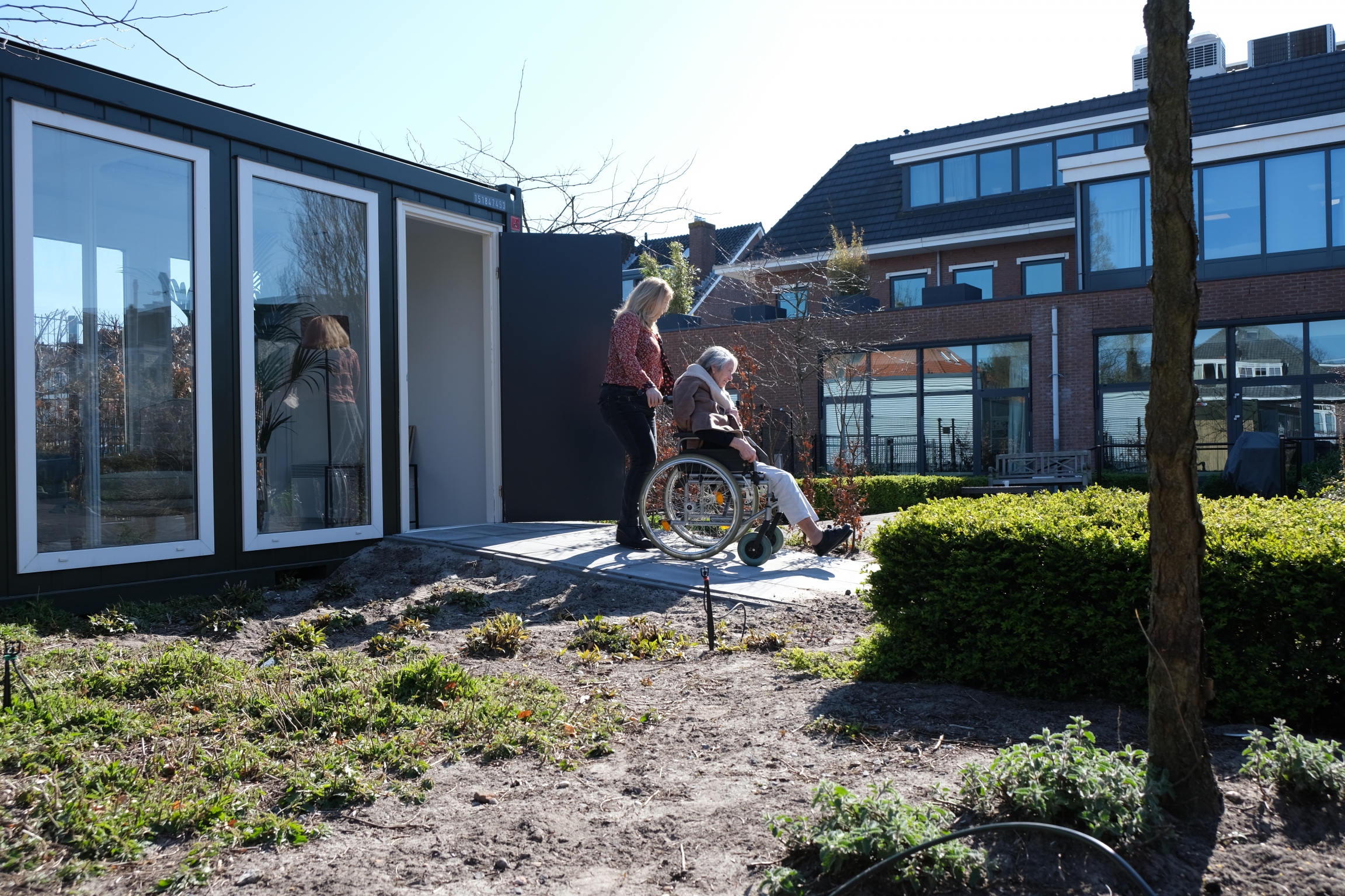 Two people exit The Glass Garden House, designed to allow family and friends to visit elders at the nursing home during the coronavirus pandemic. A woman pushes another woman in a wheelchair outside a glass garden house.