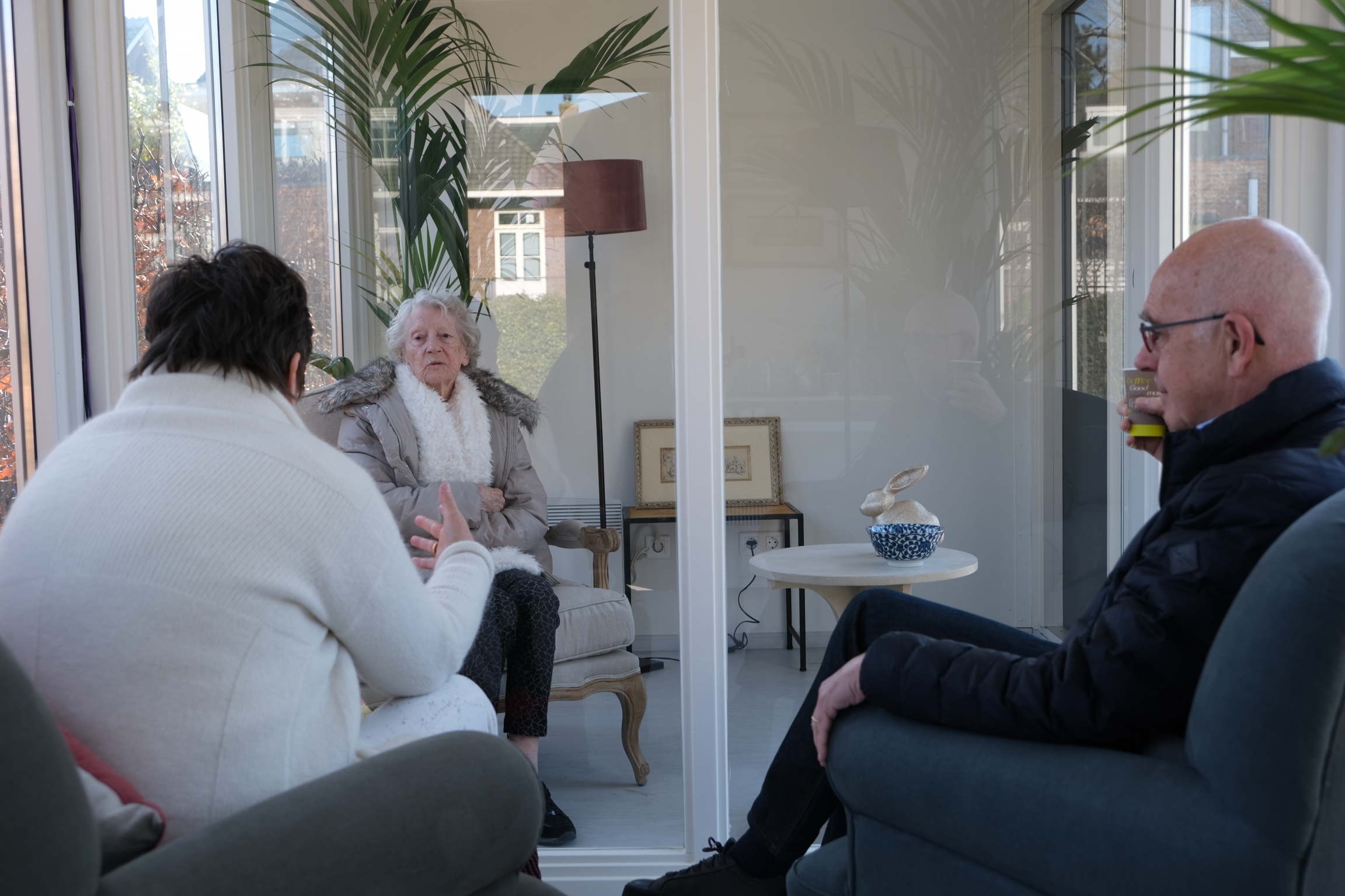 Two adults visit with an elder at The Glass Garden House. Two adults visit with an elder in a "glass cabin" separated by a pane of glass.