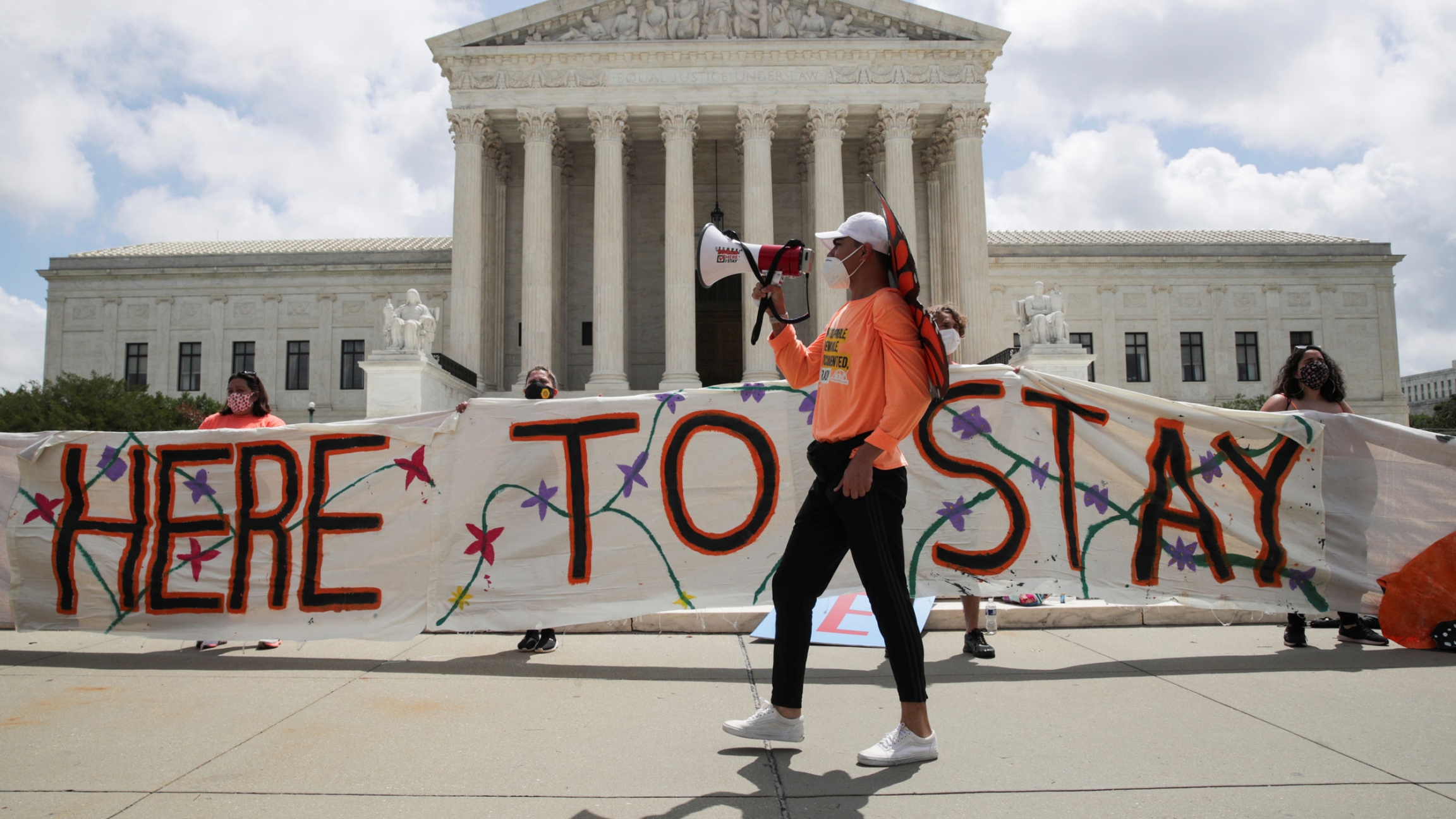 DACA recipients and their supporters celebrate outside the US Supreme Court after the court ruled in a 5-4 vote that President Donald Trump's 2017 move to rescind the Deferred Action for Childhood Arrivals program was unlawful, in Washington, DC, June 18, Several people are shown holding a large banner that says, 'Here To Stay" painted on it.