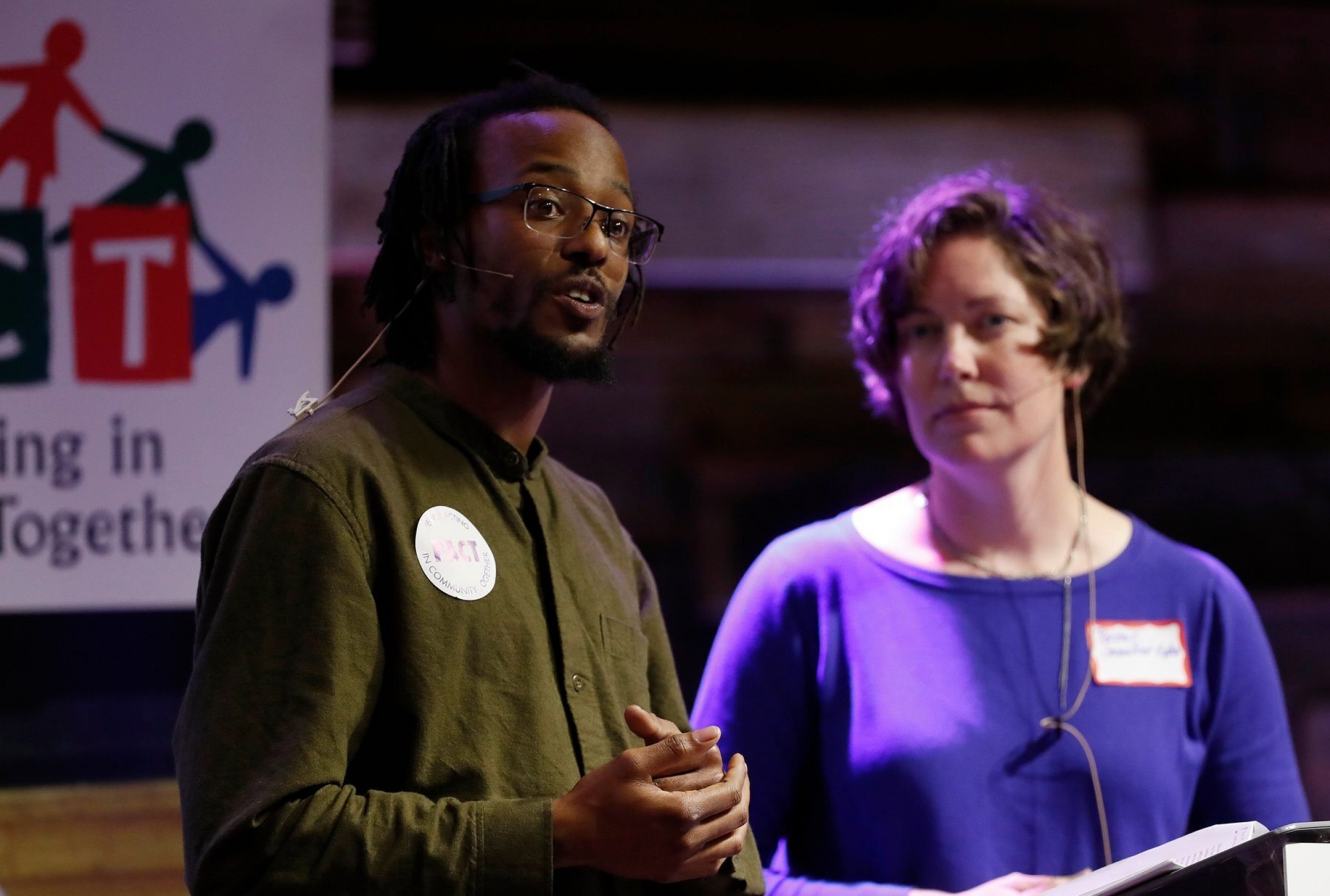 Derrick Sanderlin, left, a community organizer in San Jose, California, speaks at a meeting of People Acting in Community Together, or PACT, a group that seeks to create a more just community. A Black, male community organizer wearing a green shirt speaks next to a white woman wearing a purple shirt at a meeting of People Acting in Community Together in San Jose, California.