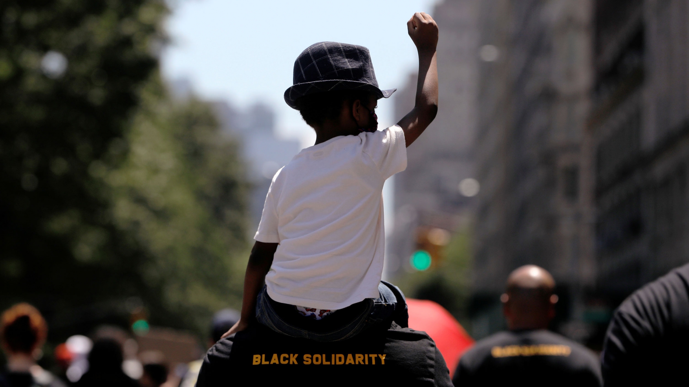 A child takes part in a rally as people march down Central Park West during events to mark Juneteenth amid nationwide protests against racial inequality, New York City, New York, June 19, 2020. A young boy is shown wearing a white t-shirt and had while sitting on the shoulder of a man wearing a shirt with "Black Solidarity" printed on it.