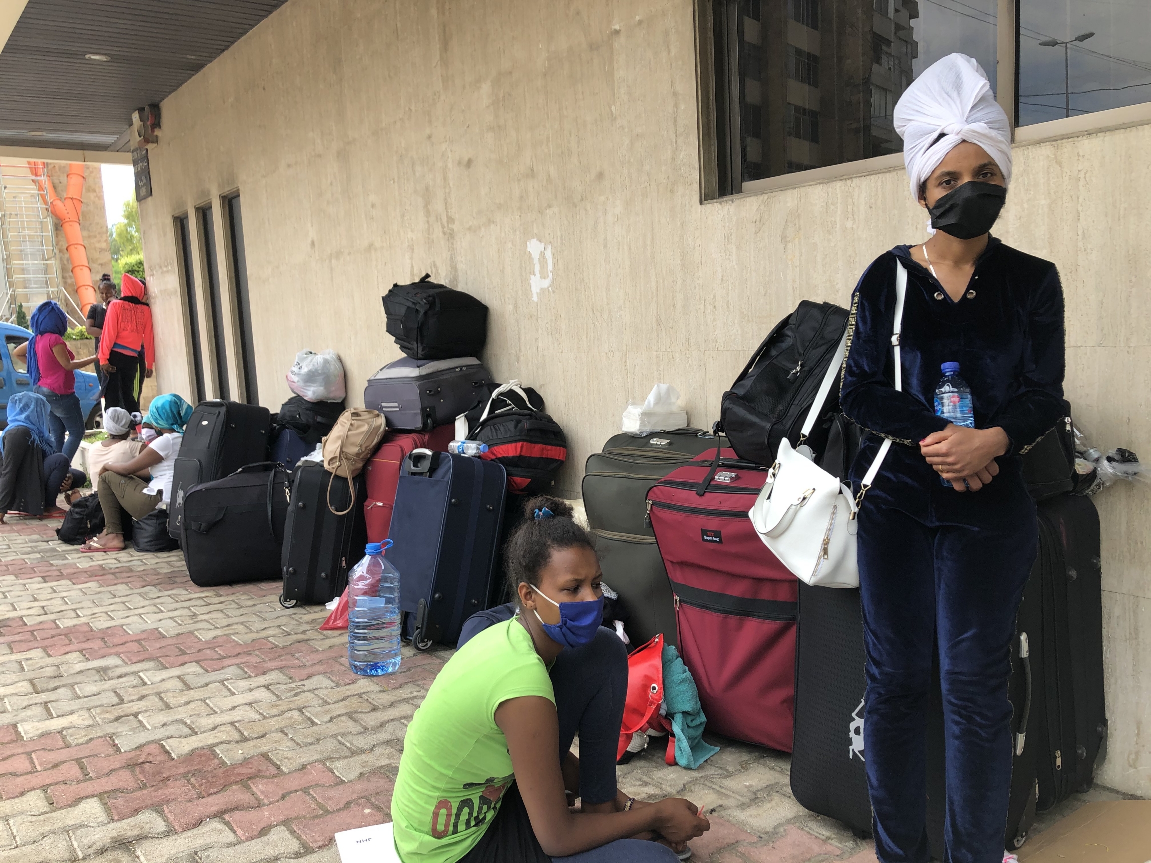 Former domestic workers from Ethiopia wait outside the Ethiopian Embassy in Beirut, Lebanon. One Ethiopian woman in a green shirt sits and one woman in a white head wrap stands outside.
