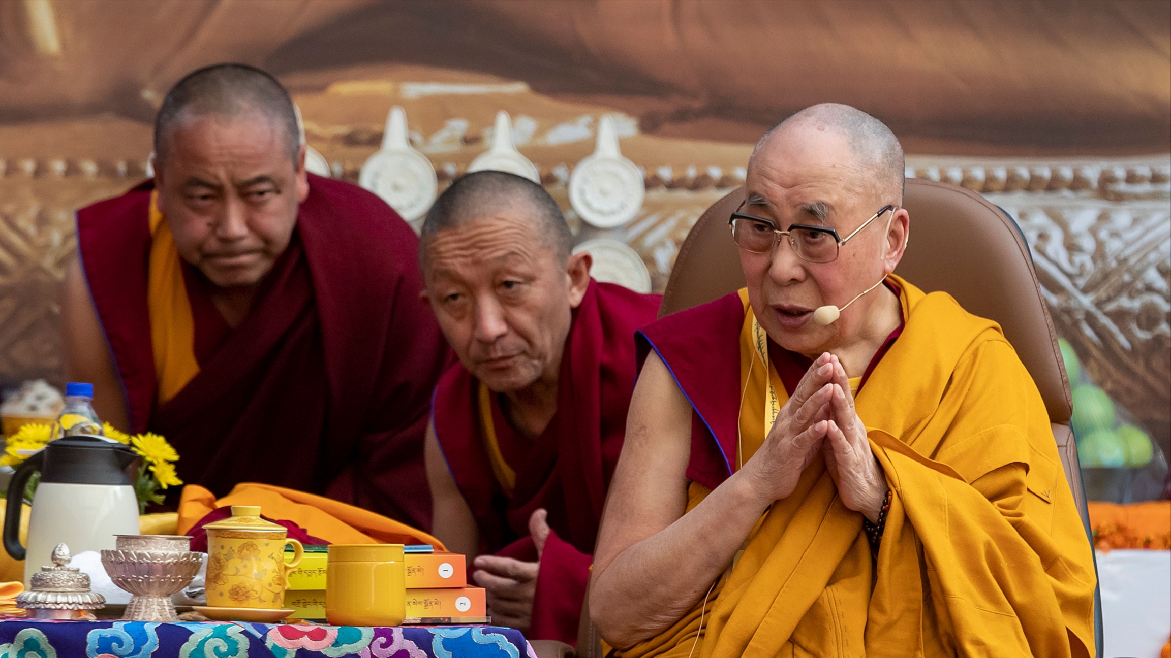 His Holiness the Dalai Lama, pictured with some of his aides, during an event at the Kirti Monastery in Dharmsala, India on Dec. 7, 2019, marking the 25th founding anniversary of the monastery. The Dalai Lama speaks while wearing a yellow robe. He is surrounded by two man on red robes in a monastery.