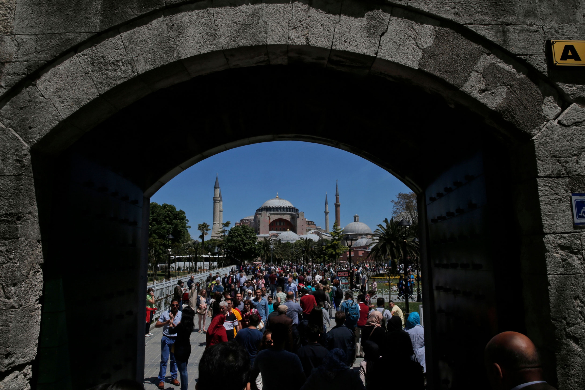 Visitors walk toward the Byzantine-era Hagia Sophia, an UNESCO World Heritage site in Istanbul, Turkey. A large crowd of people are shown through an open under a stone passageway with the iconic Hagia Sophia in the distance.