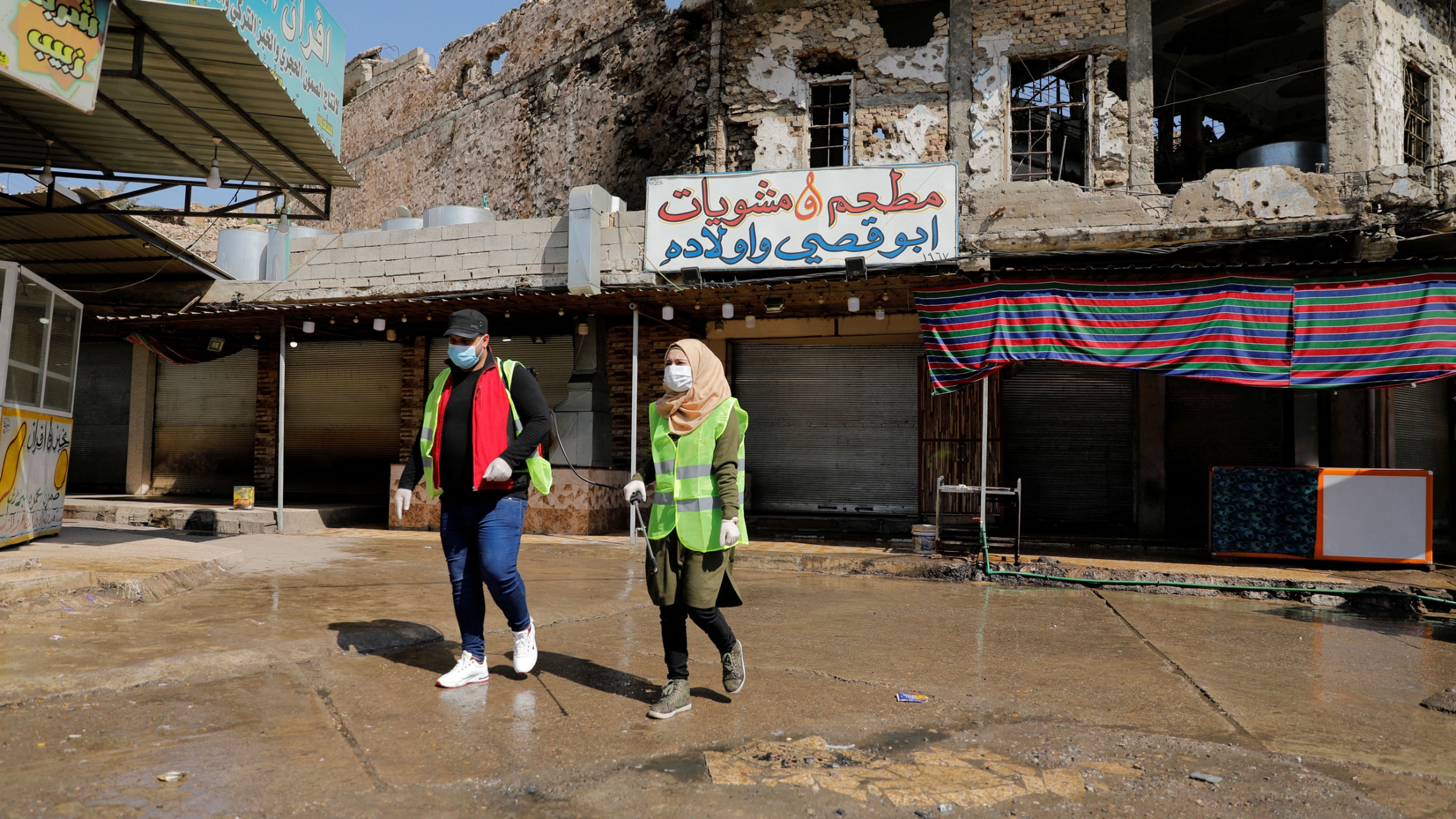 Iraqi young volunteers spray with disinfectants to combat an outbreak of coronavirus in Mosul, Iraq, March 15, 2020. Two young people are shown walking in an empty courtyard and wearing safety vests and protective face masks.