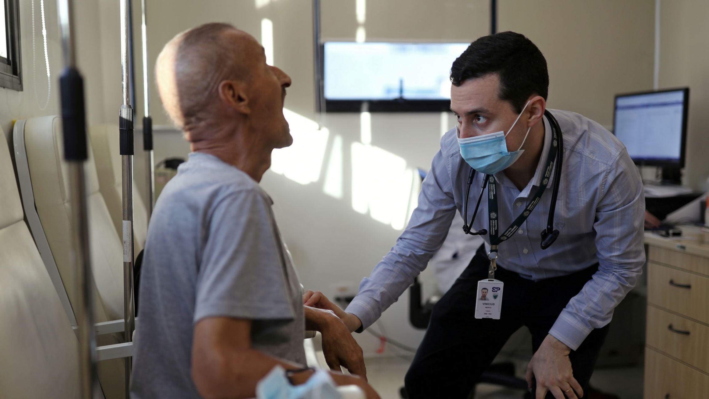 Vinicius Molla, a hematologist and volunteer of the clinical trial of Oxford University's COVID-19 vaccine, examines a patient at a consulting room in São Paulo, Brazil July 9, 2020. An older man is shown seated and with his mouth open wide with a doctor wearing a face mask looking into his mouth.