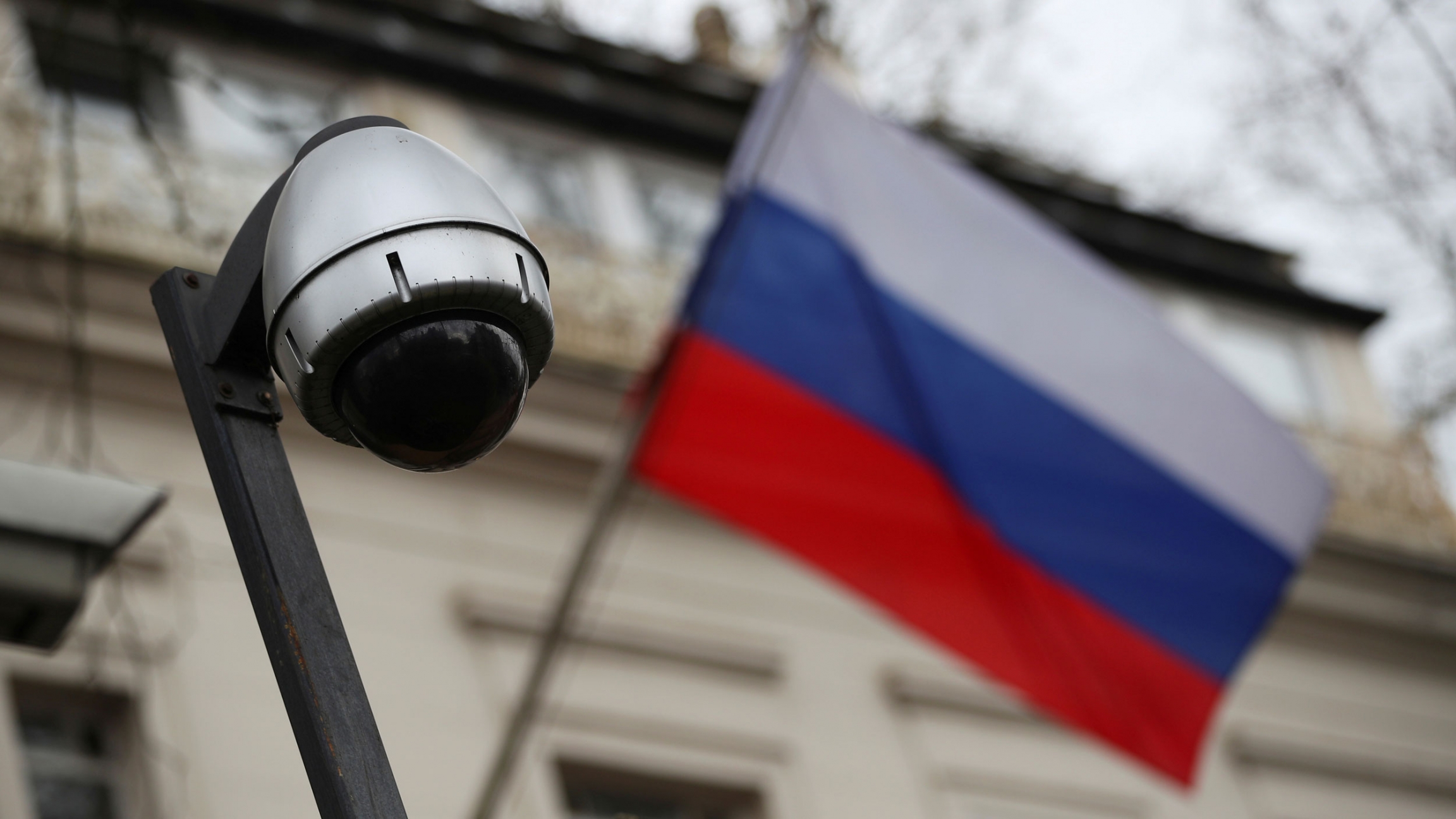 A security camera is seen with a Russian flag flying outside the consular section of Russia's embassy in London, Britain, 2018. The dark lens of a security camera is shown with a Russian Federation flag in soft focus in the background.
