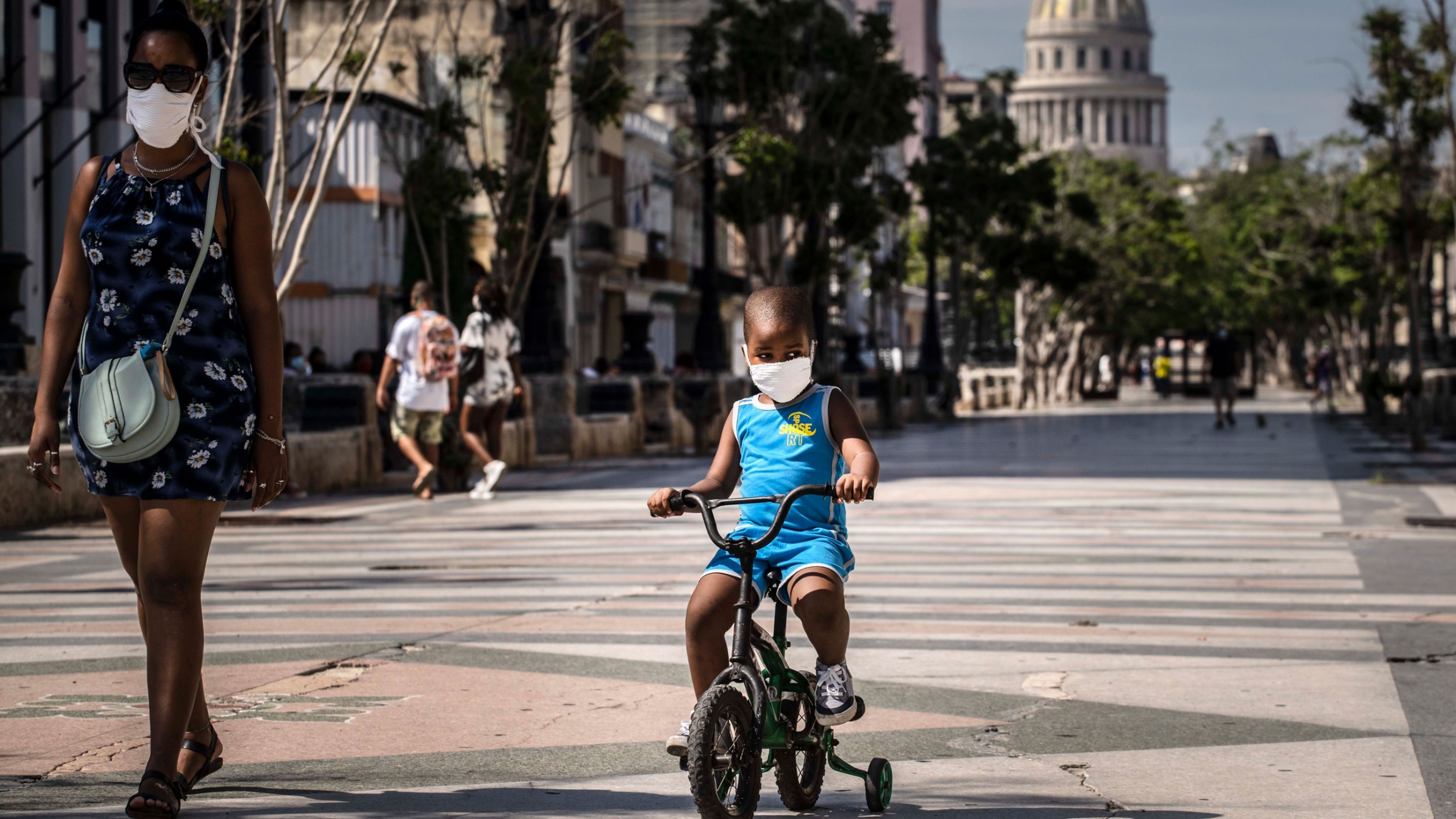 A woman accompanies a child on a bicycle, both wearing protective face masks as a precaution amid the coronavirus pandemic, in Havana, Cuba, Friday, July 3, 2020. A young child is shown wearing a face mask and a blue outfit and riding a small bicycle with training wheels.