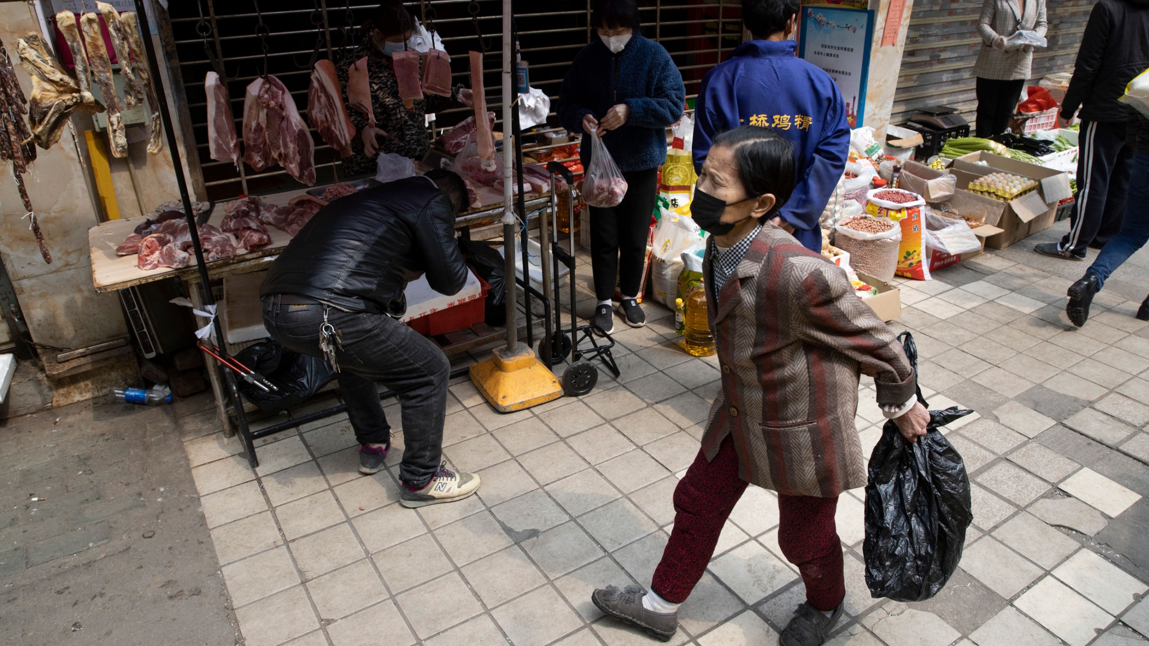 Residents walk past stalls near a partially closed off market due to the coronavirus outbreak in Wuhan in central China, April 3, 2020. Several people are shown outside of a partially closed market stall with several pieces of meat hanging in the background.