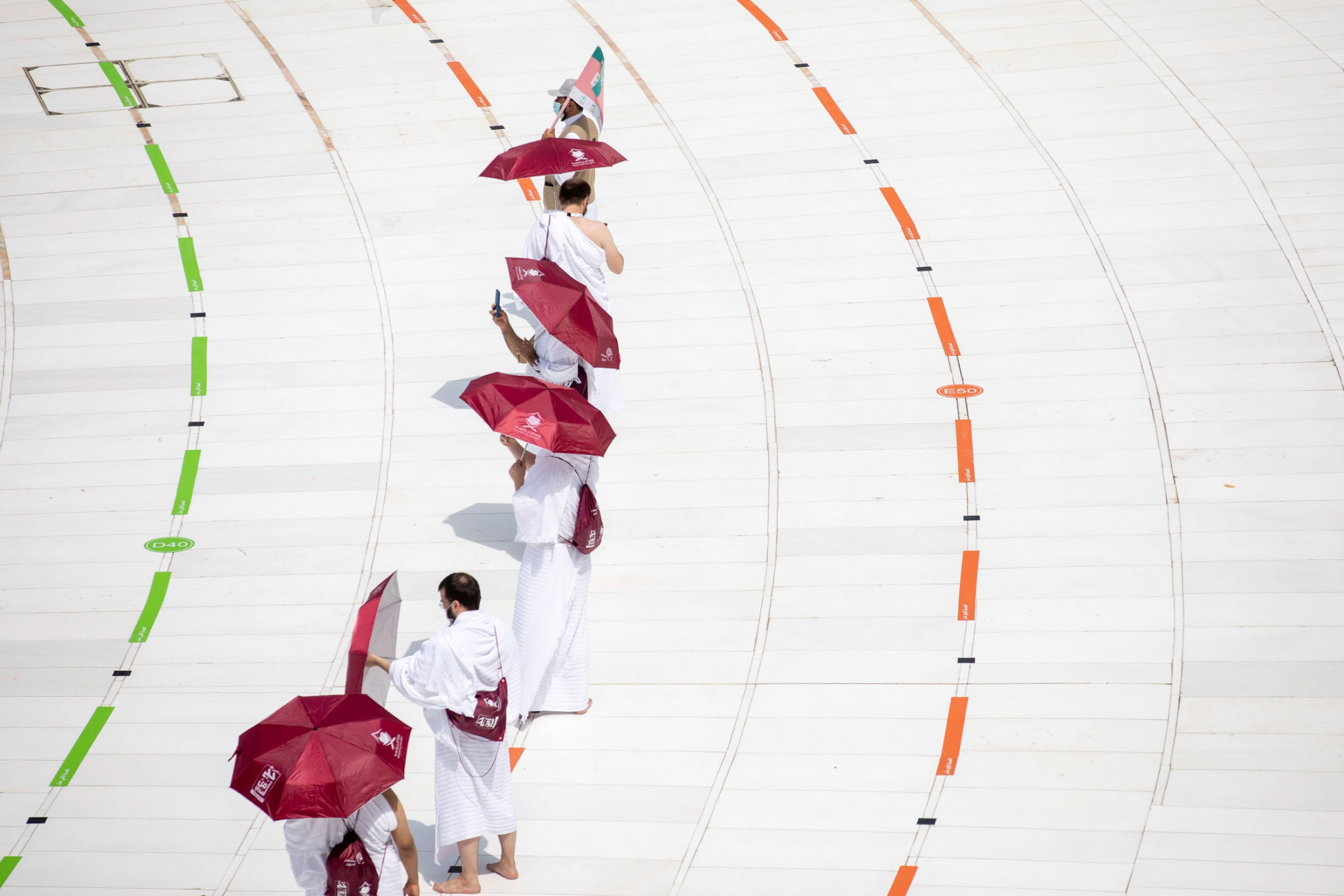 Pilgrims keep social distancing during the hajj. A line of people are shown wearing white robes and carrying red umbrellas.
