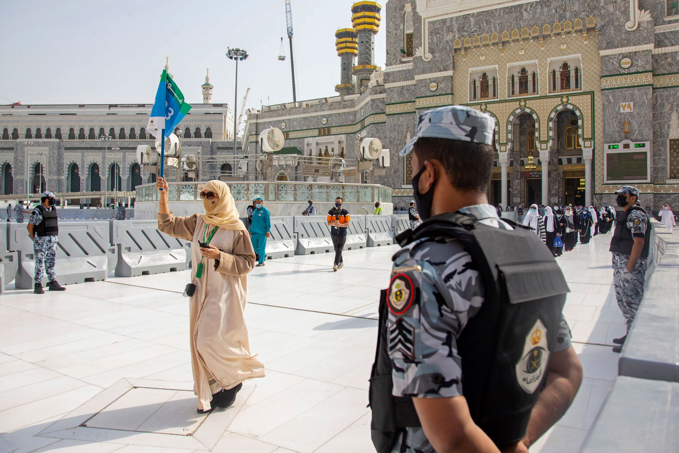 Security personnel standby as a limited numbers of pilgrims arrive at the Grand Mosque. A security official is shown in the near ground with a woman in the distance walking and holding up a guide flag.