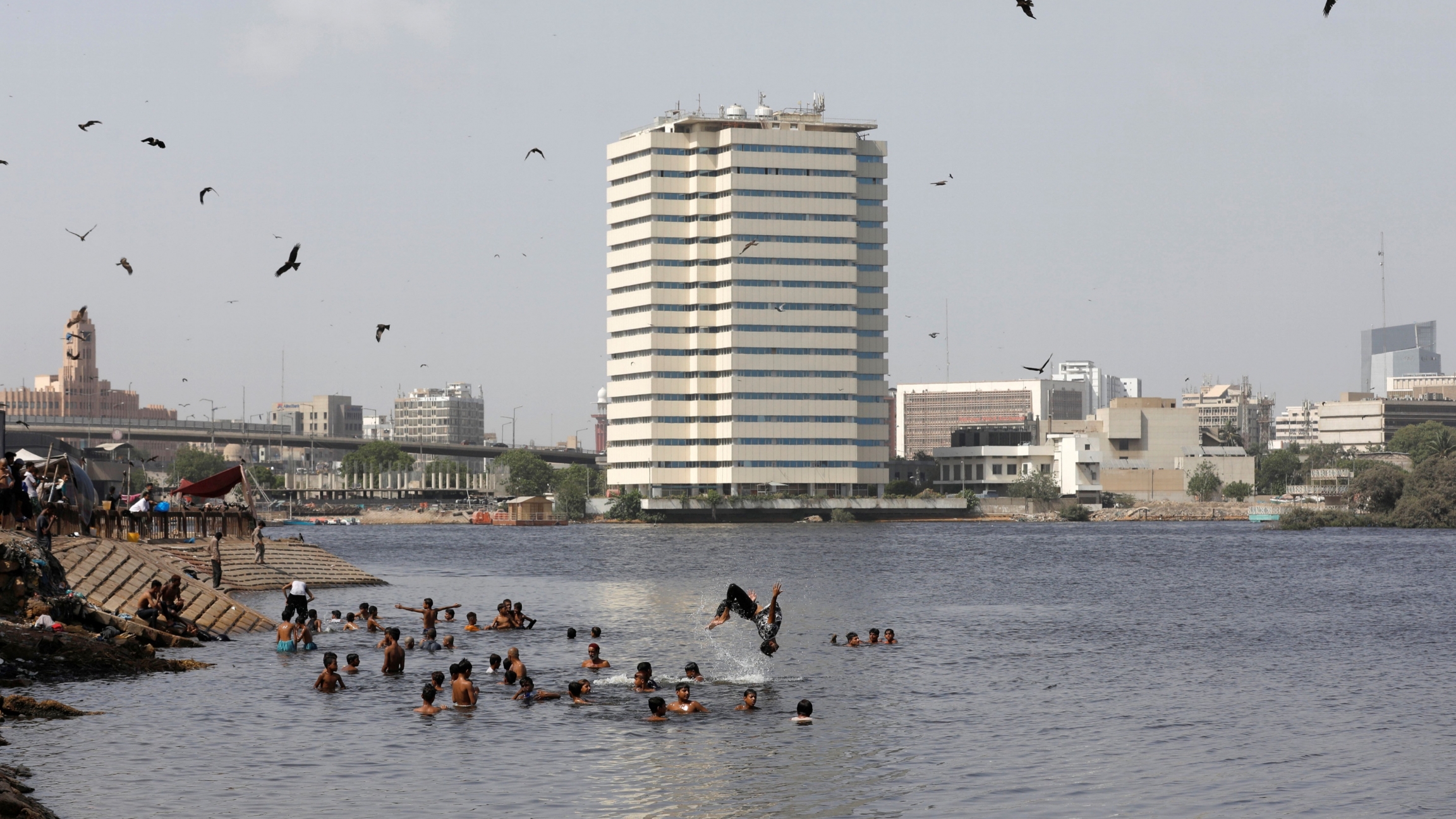 Men and children take bath as they cool off during a hot and a humid day at China Creek area, Karachi, Pakistan June 8, 2020. Adults and children swim in water near a large, white building in the background and birds flying overhead