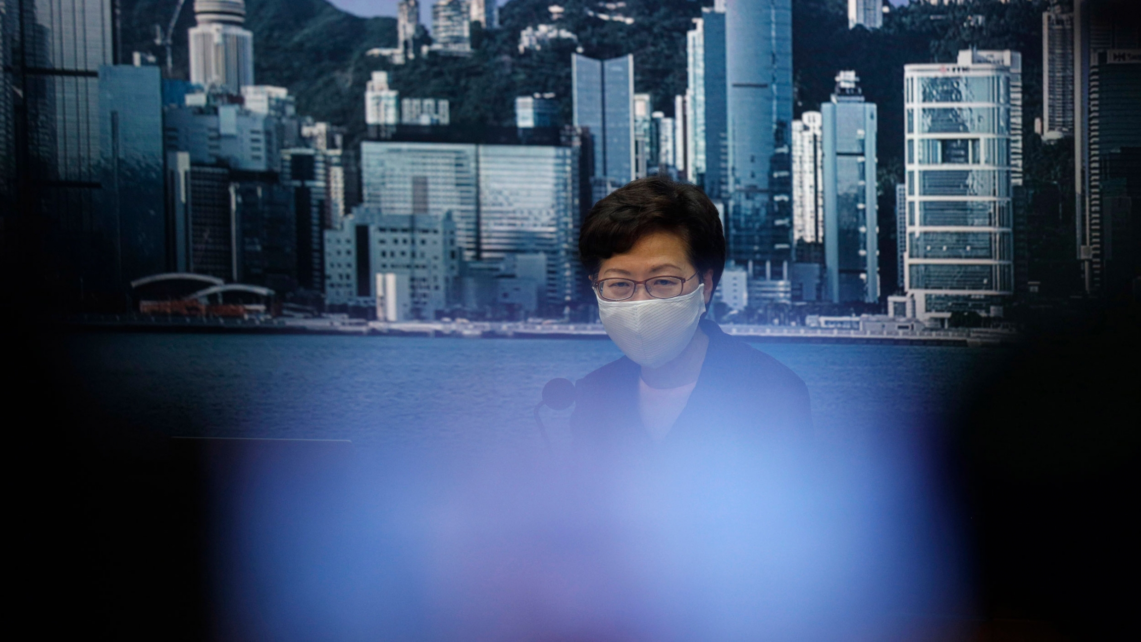 Hong Kong Chief Executive Carrie Lam arrives for a news conference in Hong Kong, Friday, July 31, 2020. Hong Kong leader Lam announced that the government will postpone highly anticipated legislative elections by one year. Hong Kong Chief Executive Carrie Lam is shown wearing a face mask with an image of the Hong Kong coastline in the background.