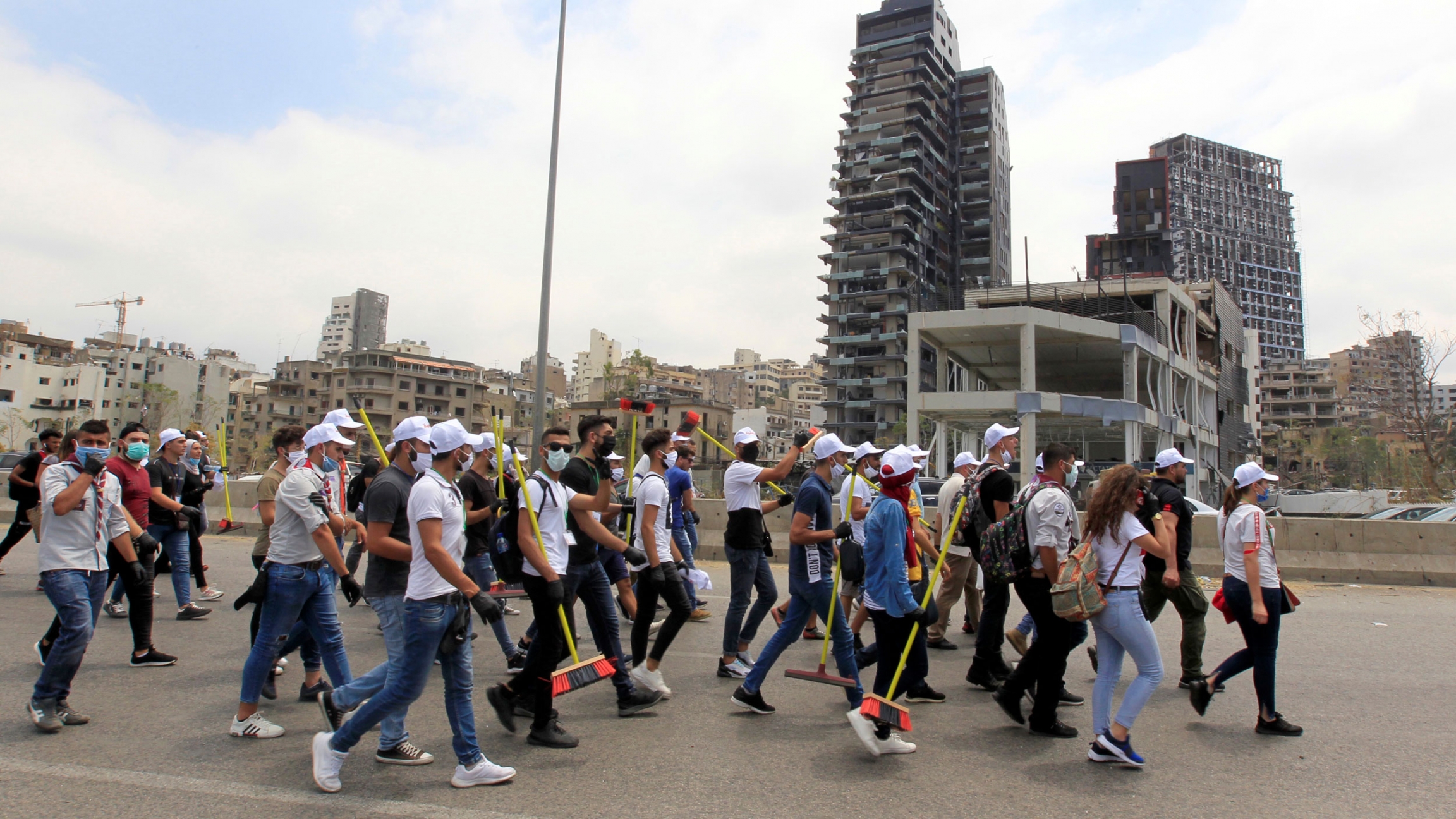 People walk as they clean near site of Tuesday's blast in Beirut's port area, Lebanon, August 6, 2020. A crowd of people are shown walking in a street with many carrying a broom.