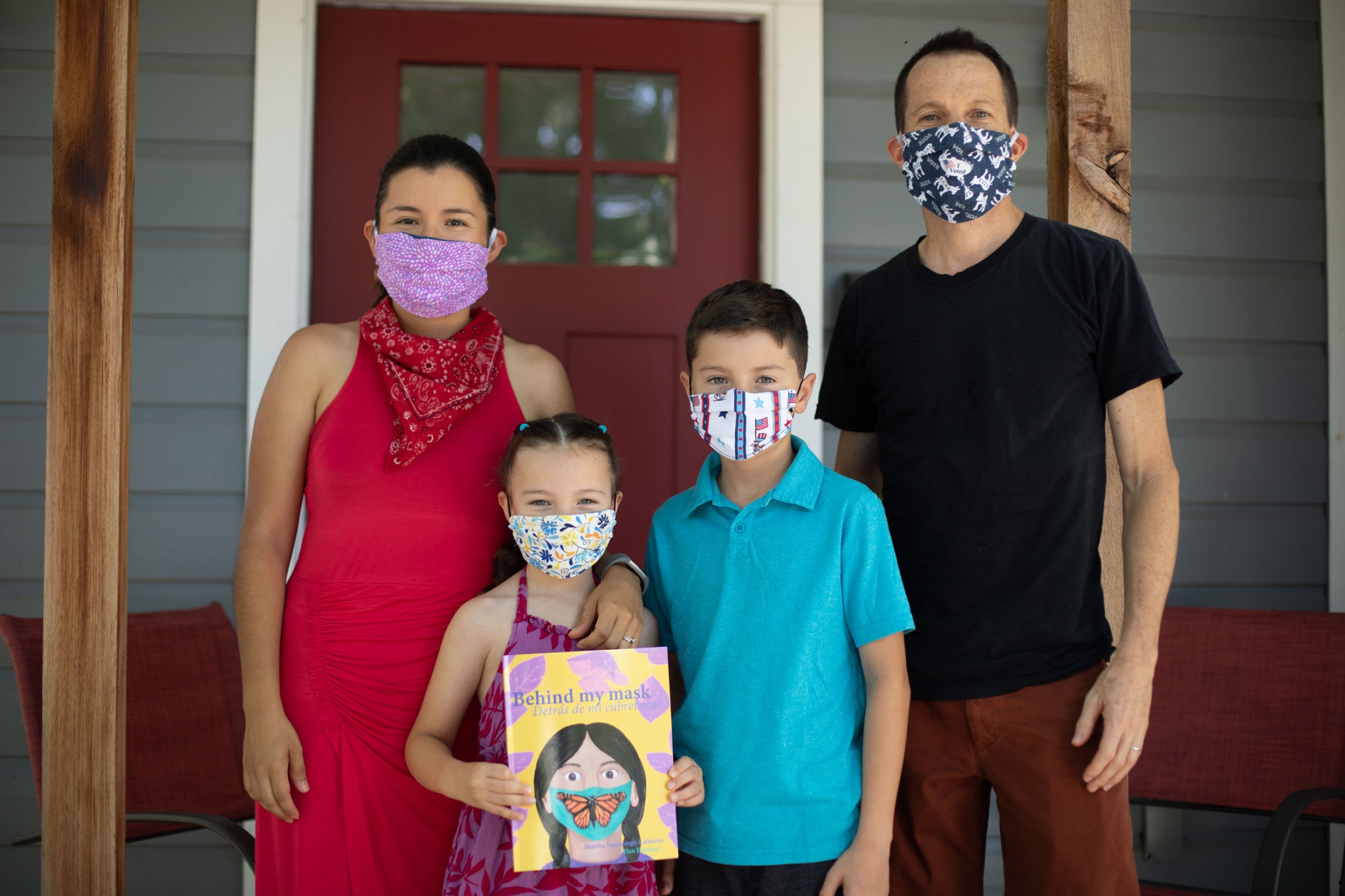 Martha Samaniego Calderón and Dan Heiman pose for a portrait with their children Natalia and Nicolas, at their home. A family of four pose for a photo wearing masks. The young girl holds a book in her hands.