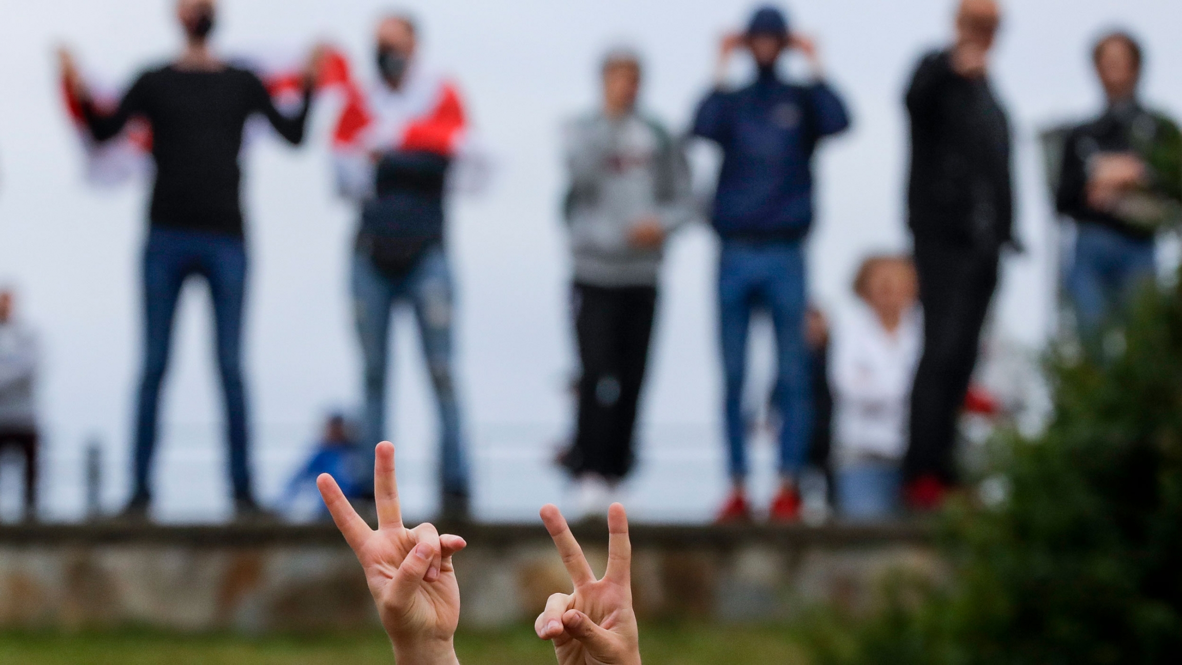 Protesters gather on a street against election results in Minsk, Russia, Aug. 12, 2020. A pair of hands are shown in the nearground flashing peace signs with several people standing in a row in the background.