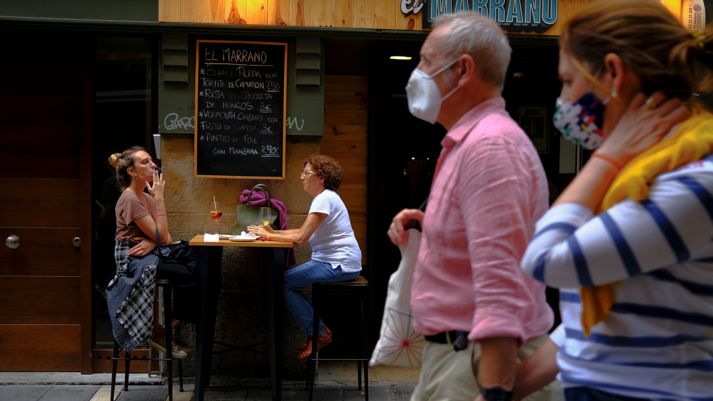 Two women smoke cigarettes in a bar while pedestrians wearing face walk past, in Pamplona, northern Spain, Aug. 17, 2020. Spanish authorities have announced new restrictions, one of them about smoking in public places, to prevent the COVID-19. A couple is shown in soft focus walking and wearing face masks with two women shown in the distance sitting at a cafe table and one woman is smoking a cigarette.