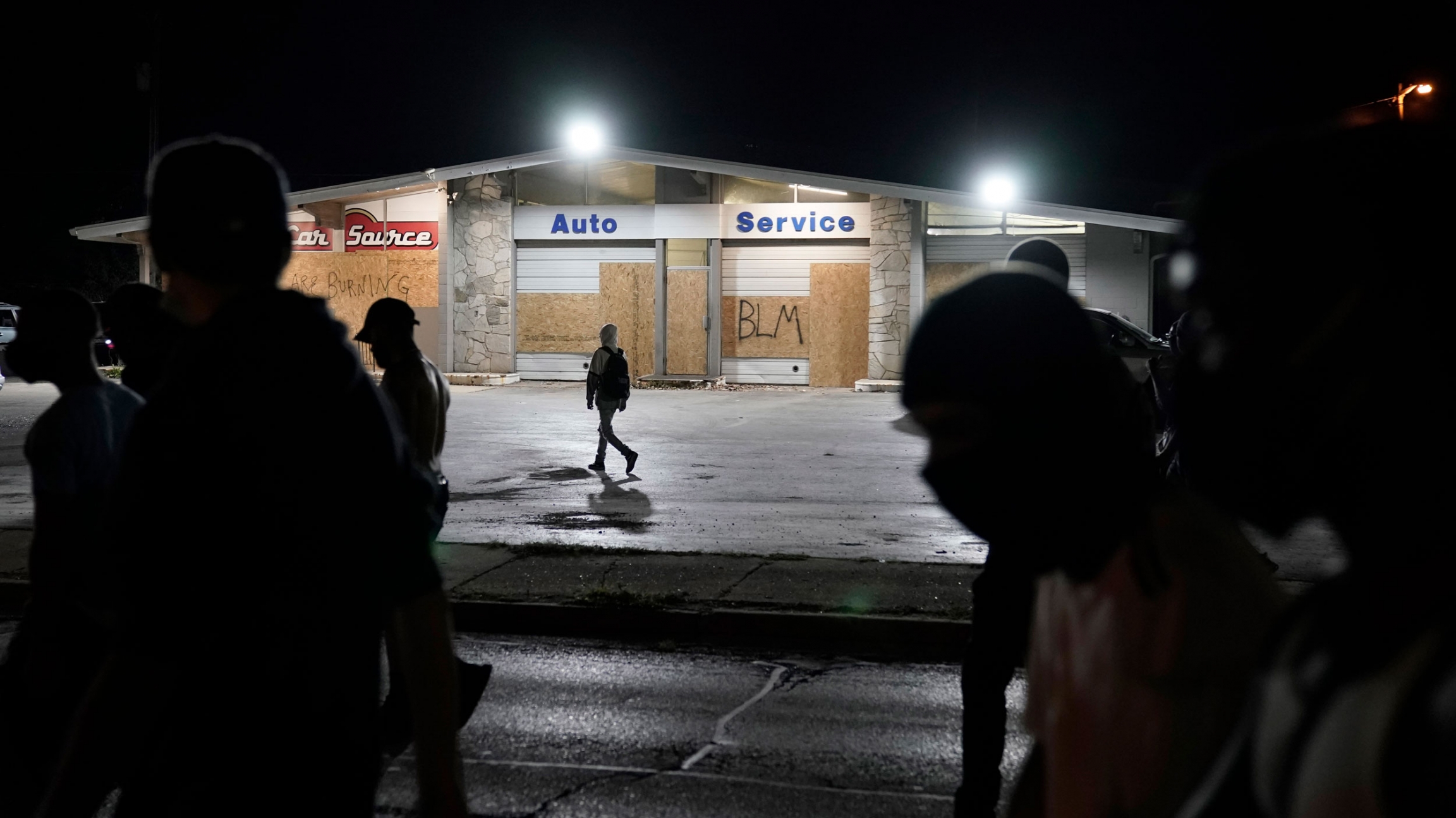 Protesters march to the site of a shooting in Kenosha, WI, Aug. 26, 2020. A white, 17-year-old was arrested after two people were shot to death during a third night of protests in Kenosha over the police shooting of a Black man, Jacob Blake. Several people are shown in shadow with a automobile service center stands in the background with boarded up windows.