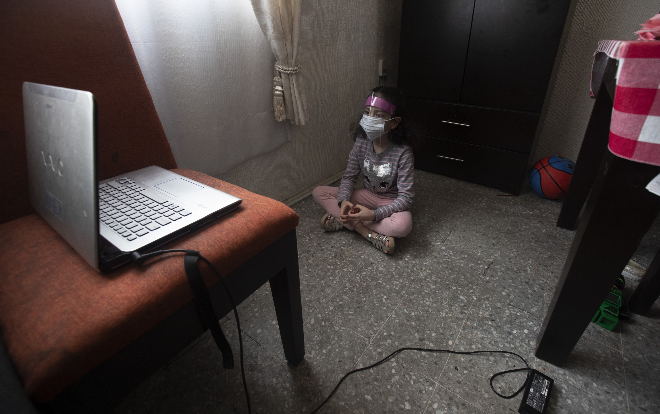 Monserrat Medina Zentella attends school via the internet, amid the new coronavirus pandemic, from her home in Mexico City, Monday, Aug. 24, 2020. A girl wears a plastic guard indoors as she watches a laptop screen.