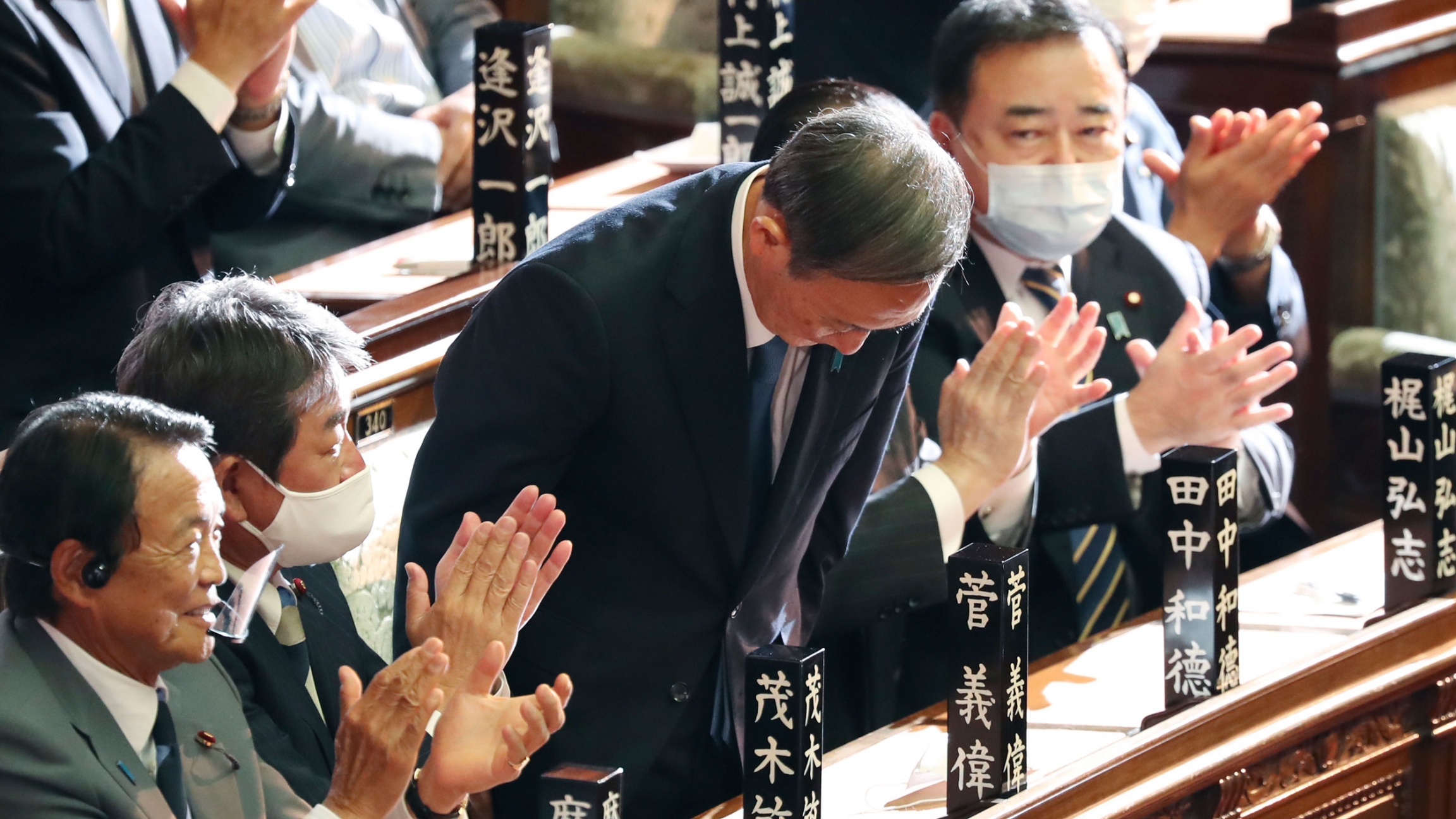 Yoshihide Suga bows after being elected as Japan's new prime minister at the parliament's lower house in Tokyo, Sept. 16, 2020. Yoshihide Suga is shown bowing and surrounded by parliamentarians on either side clapping their hands.