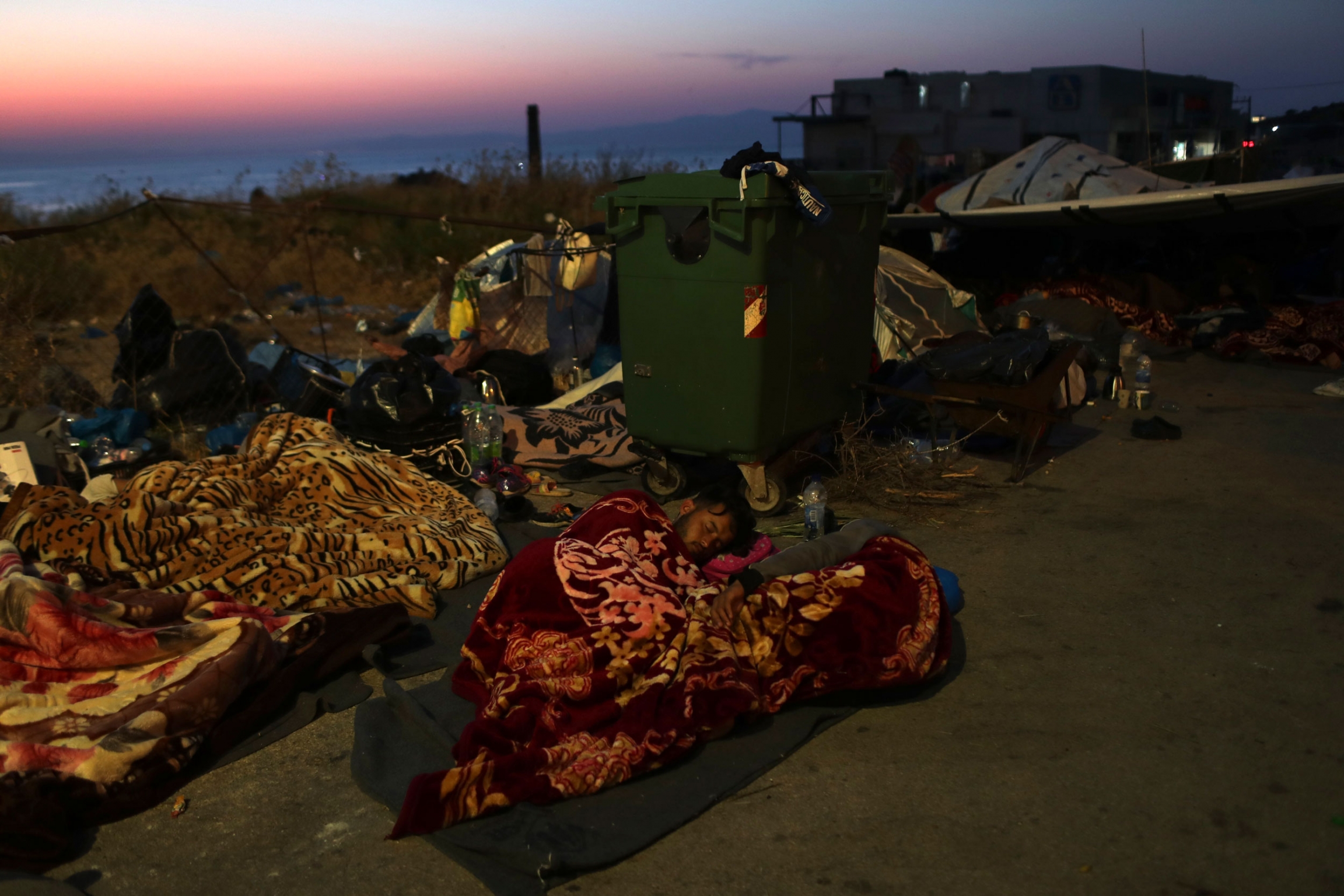 Migrants sleep next to a trash bin as they remained camped out on a road leading from Moria, on the northeastern island of Lesbos, Greece, Sept. 17, 2020. Several migrants and refugees are shown sleeping under blankets on the side of a road with a green trash bin with wheels nearby.