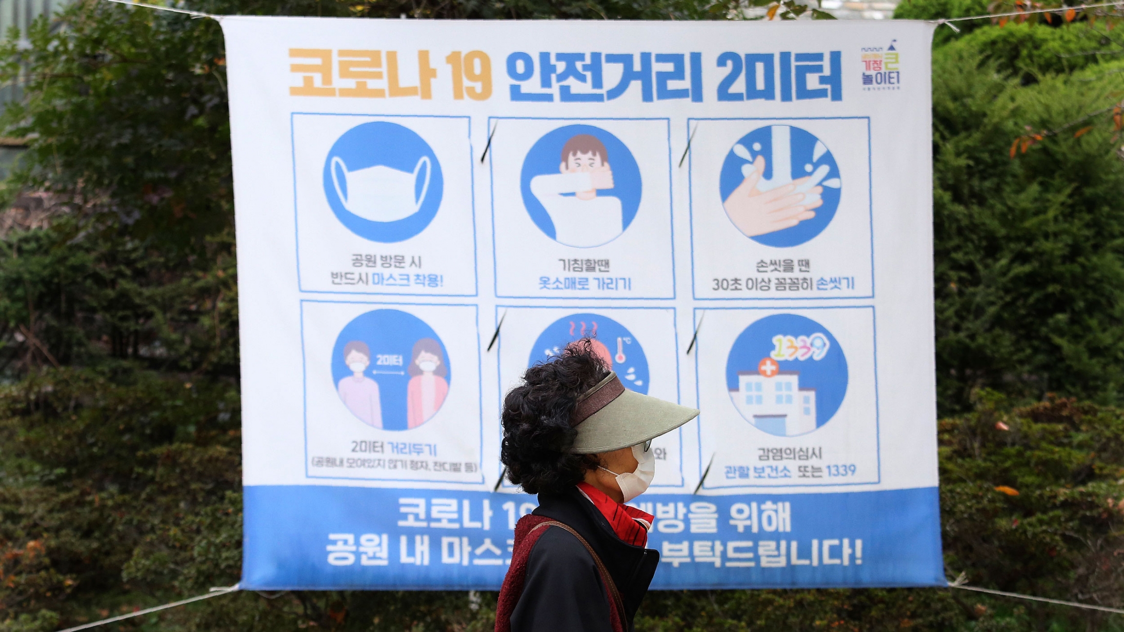 A woman wearing a face mask walks by a social distancing sign at a park in Seoul, South Korea, Oct. 16, 2020. A woman is shown wearing a visor and face mask while walking past a poster with guidance on stopping the spread of the coronavirus.