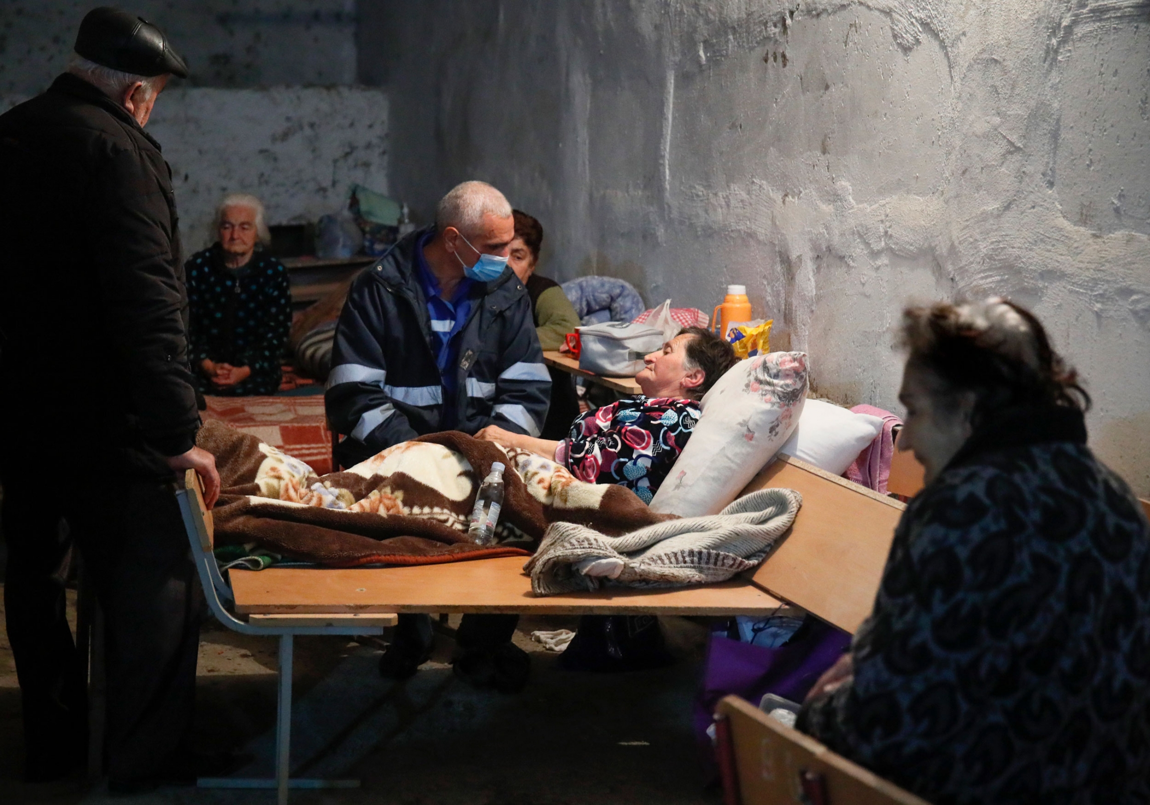 A medical worker talks to a sick woman in a bomb shelter in Stepanakert, the separatist region of Nagorno-Karabakh, Oct. 22, 2020. An man is shown sitting at the bedside of an older woman laying with her back on a large pillow.