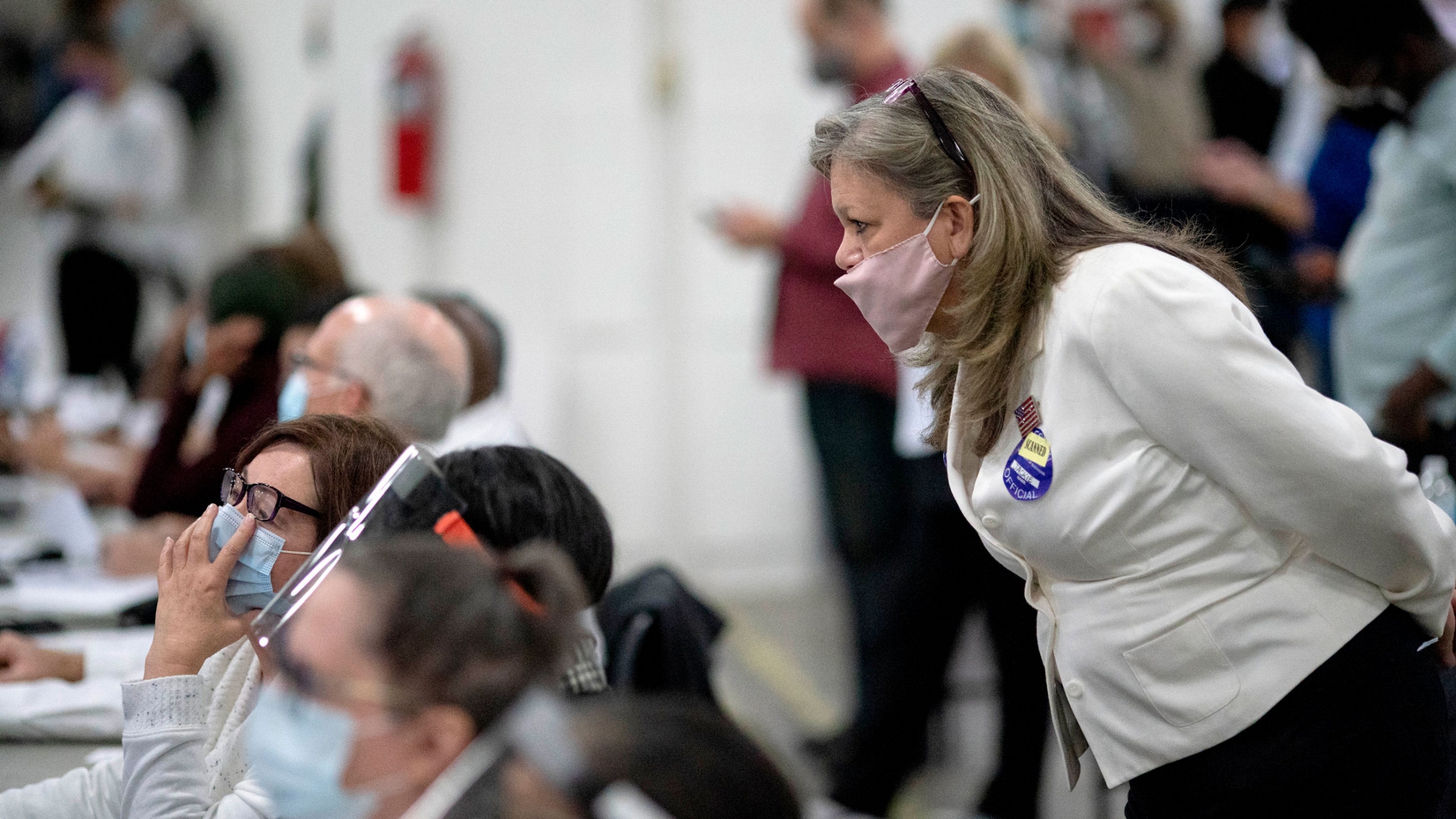 An election challenger at right watches over election inspectors as they examine a ballot as votes are counted into the early morning hours in Detroit, Michigan, Nov. 4, 2020. A woman wearing a white blazer and face mask is shown looking over the shoulder of a row of seated election inspectors.