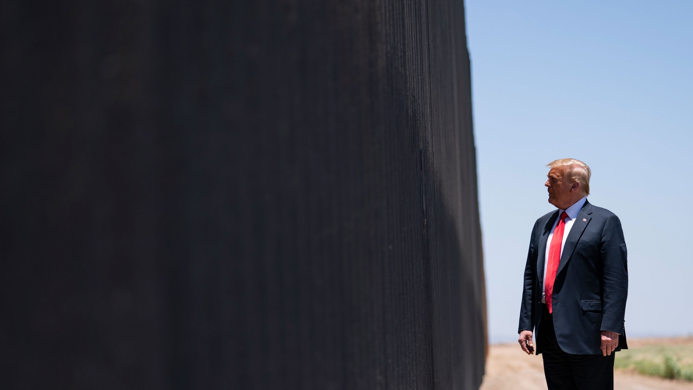 President Donald Trump tours a section of the southern border wall between the US and Mexico in San Luis, Ariz. June 23, 2020. President Donald Trump is shown wearing a blue suit and red tie while looking at the dark facade of the US-Mexico border wall.