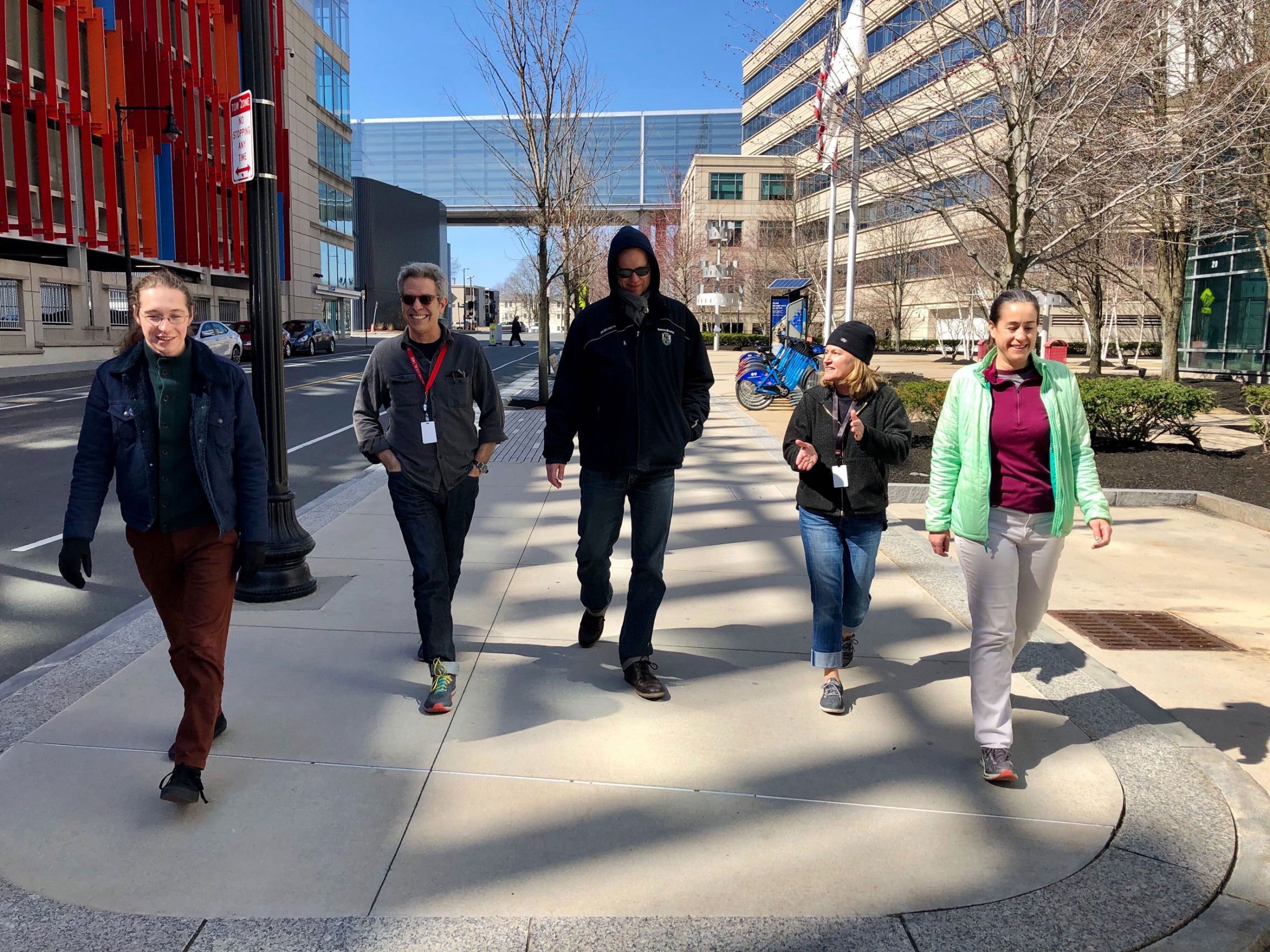 Several staff from The World's team take a break outside of GBH amid the coronavirus pandemic. Five staff members from The World are shown walking on the sidewalk next to a colorful parking garage and the GBH headquarters building, west.