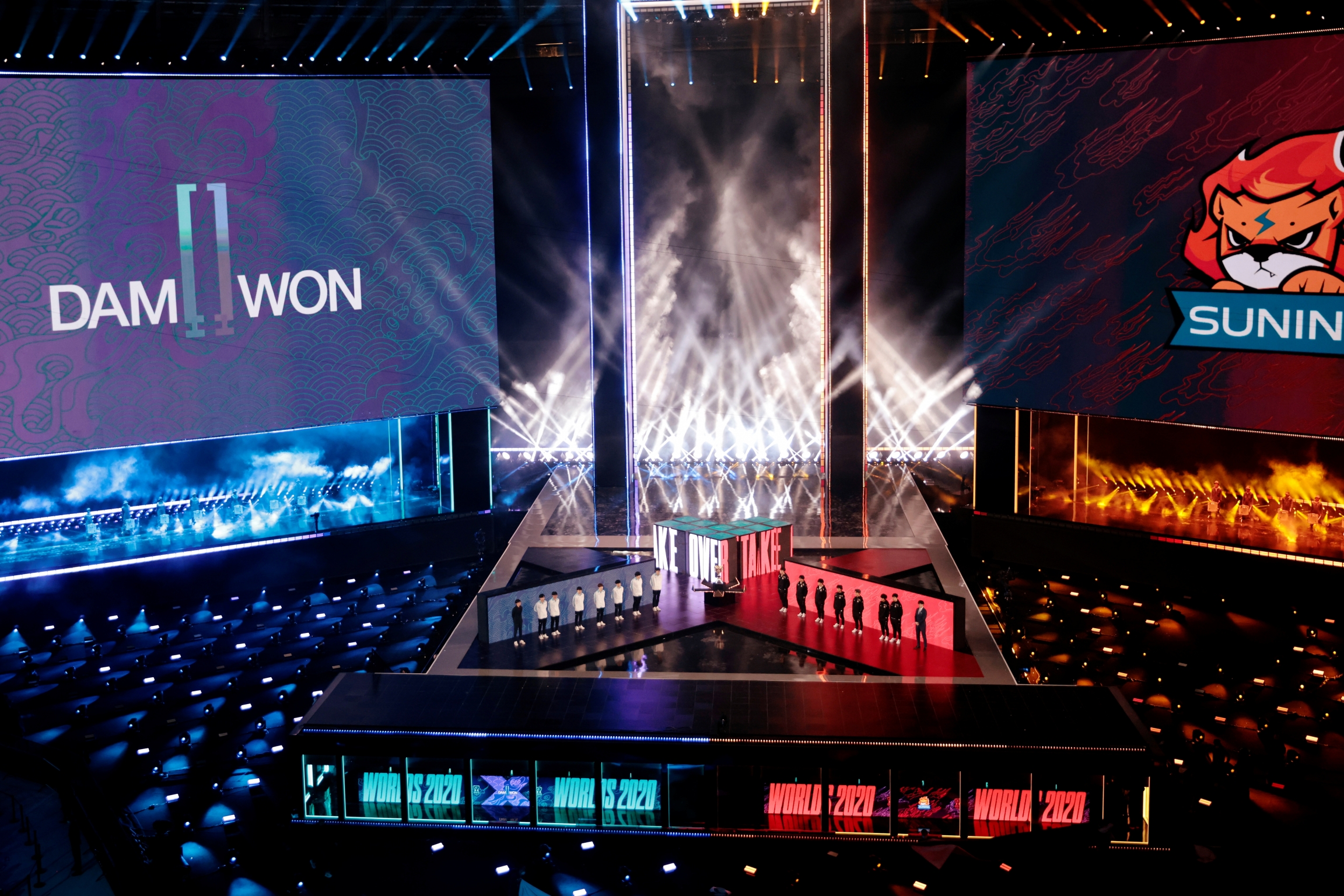 A huge stadium with blue, white and red lights features competitive esports players on stage. 