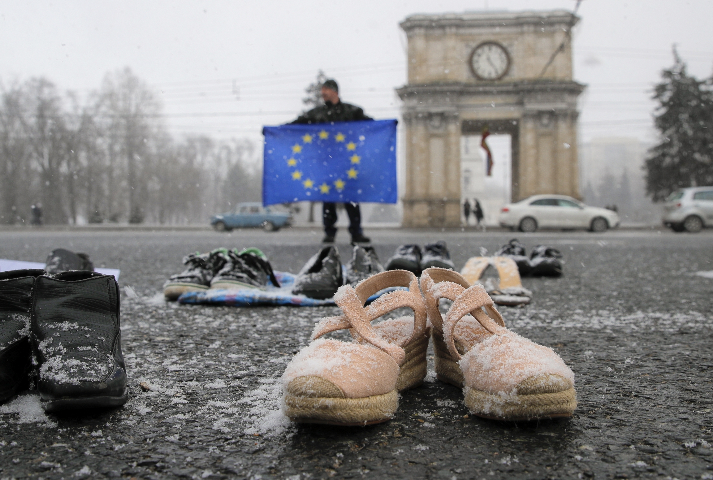 In this Saturday, Feb. 23, 2019 file photo, a man holds a European Union flag as people stand by shoes of Moldovan citizens working abroad are placed on the pavement in Chisinau, Moldova. A few pairs of shoes with snow on them lie in a square near a man with an EU flag with blue and yellow colors.